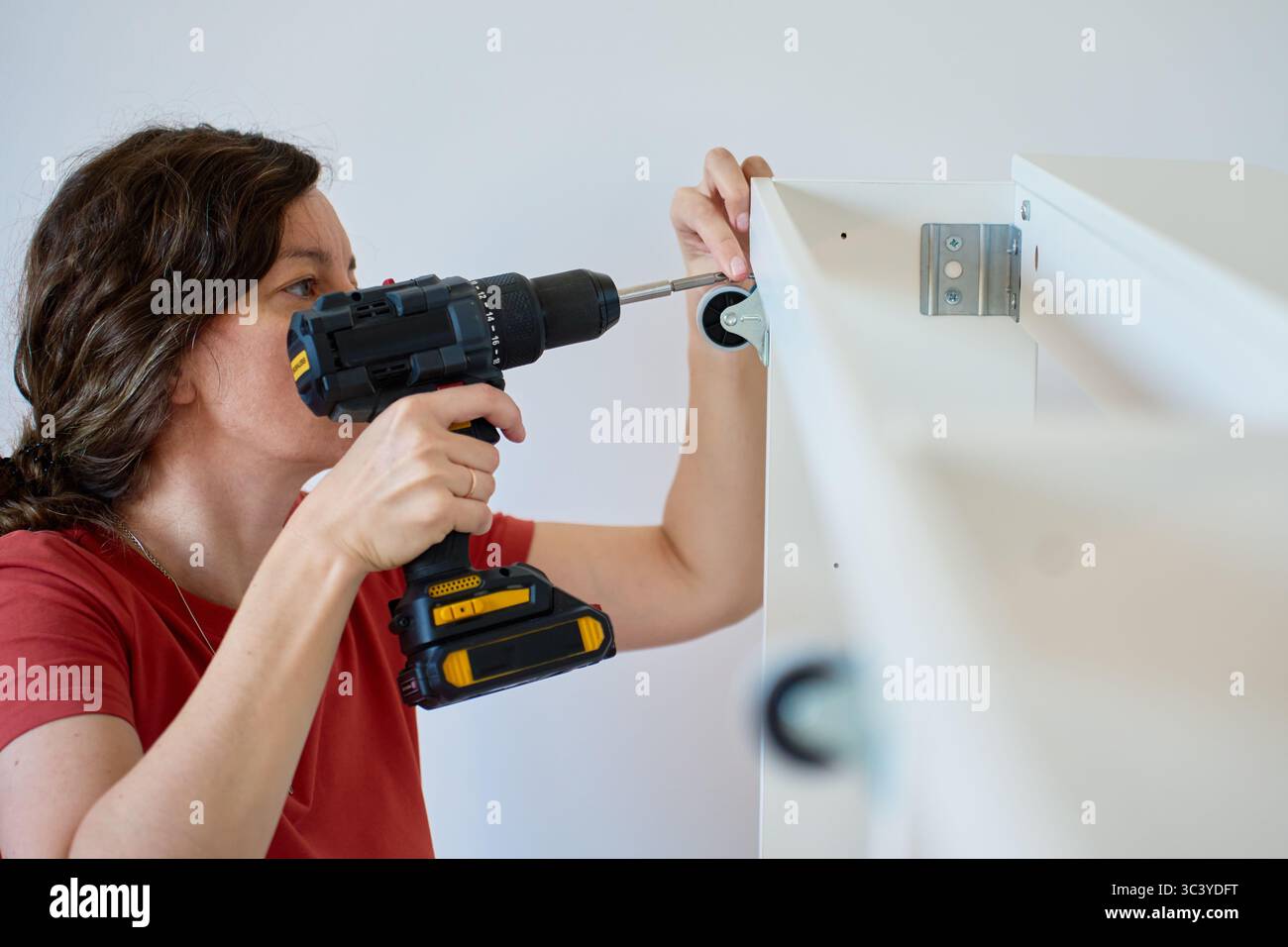 Woman using cordless drill to screw part into white bed frame. Female assembling furniture in new apartment. DIY home renovation and home organization Stock Photo