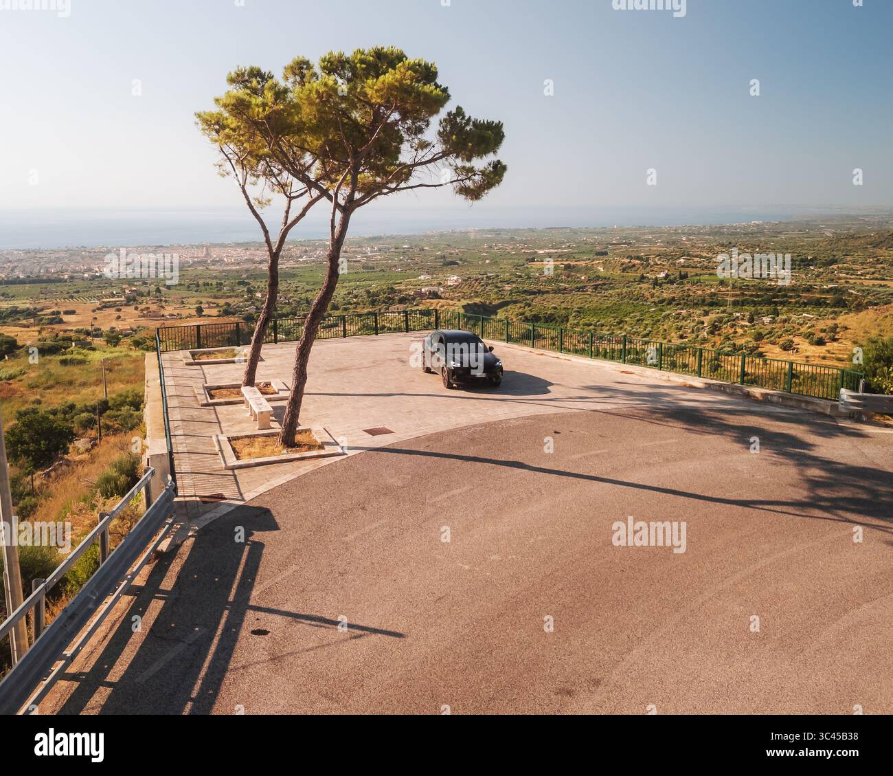 Aerial view of a dark car parked near the edge of a cliff, overlooking a valley with scattered buildings and a distant coastline, Ispica, Sicily, Ital Stock Photo
