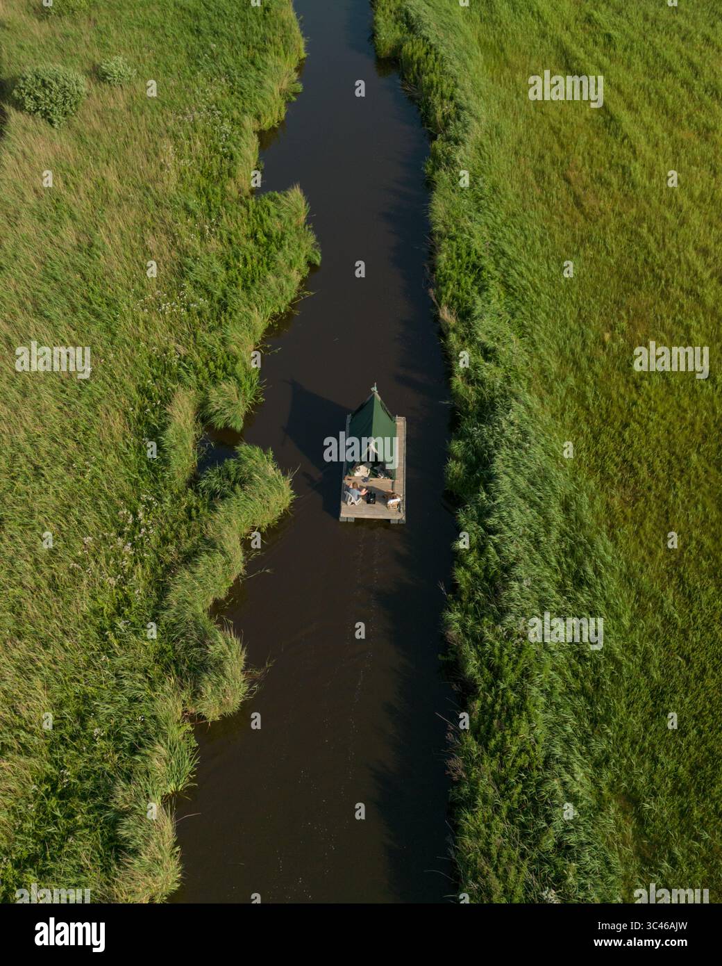 Aerial view of a solitary green-roofed structure sits serenely on a narrow waterway, flanked by vibrant green reeds, reflecting the tranquil Dutch lan Stock Photo