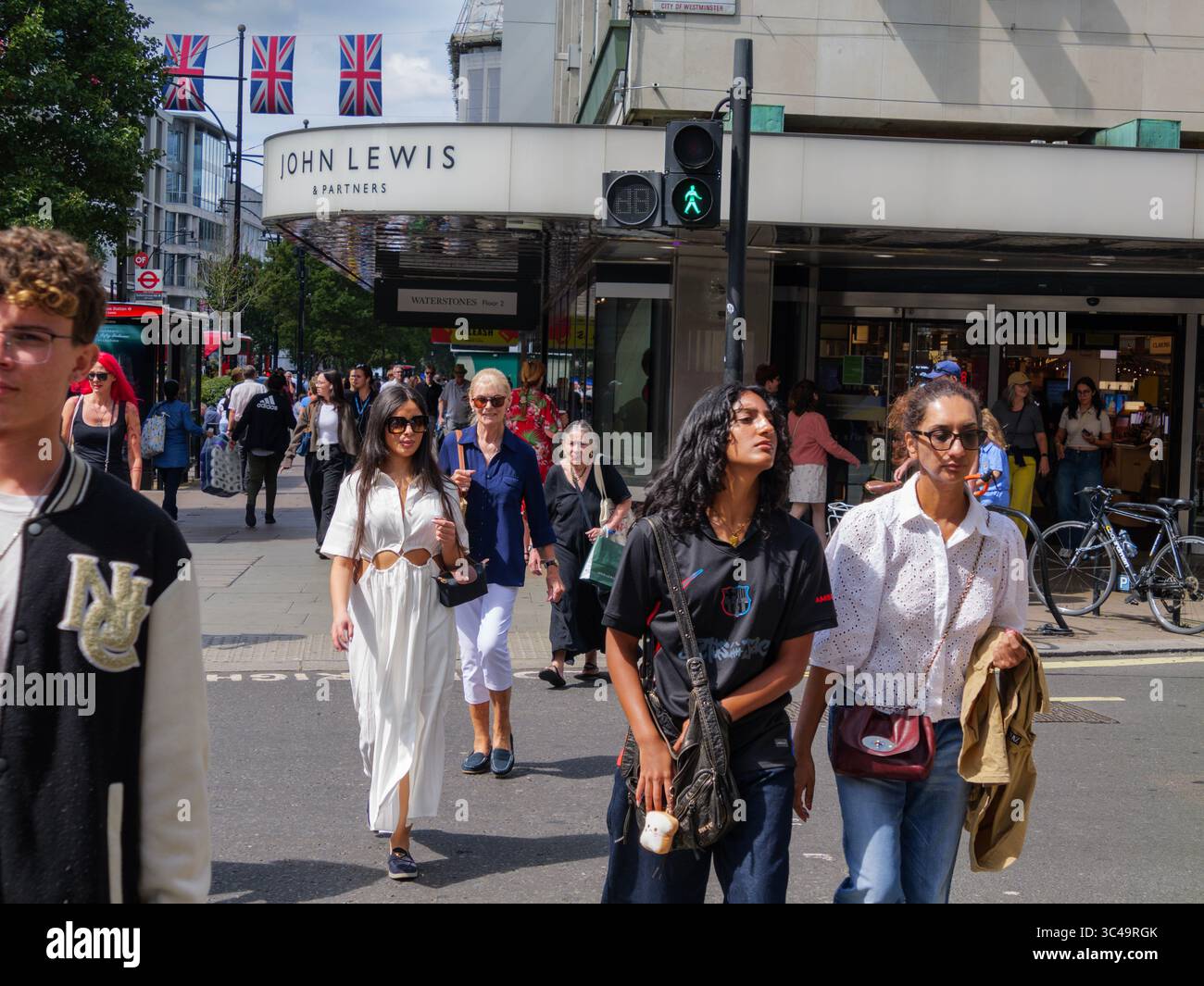 Shoppers walk past the John Lewis department store on Oxford Street, London, UK, with Union Jack flags flying above the bustling high street. Stock Photo