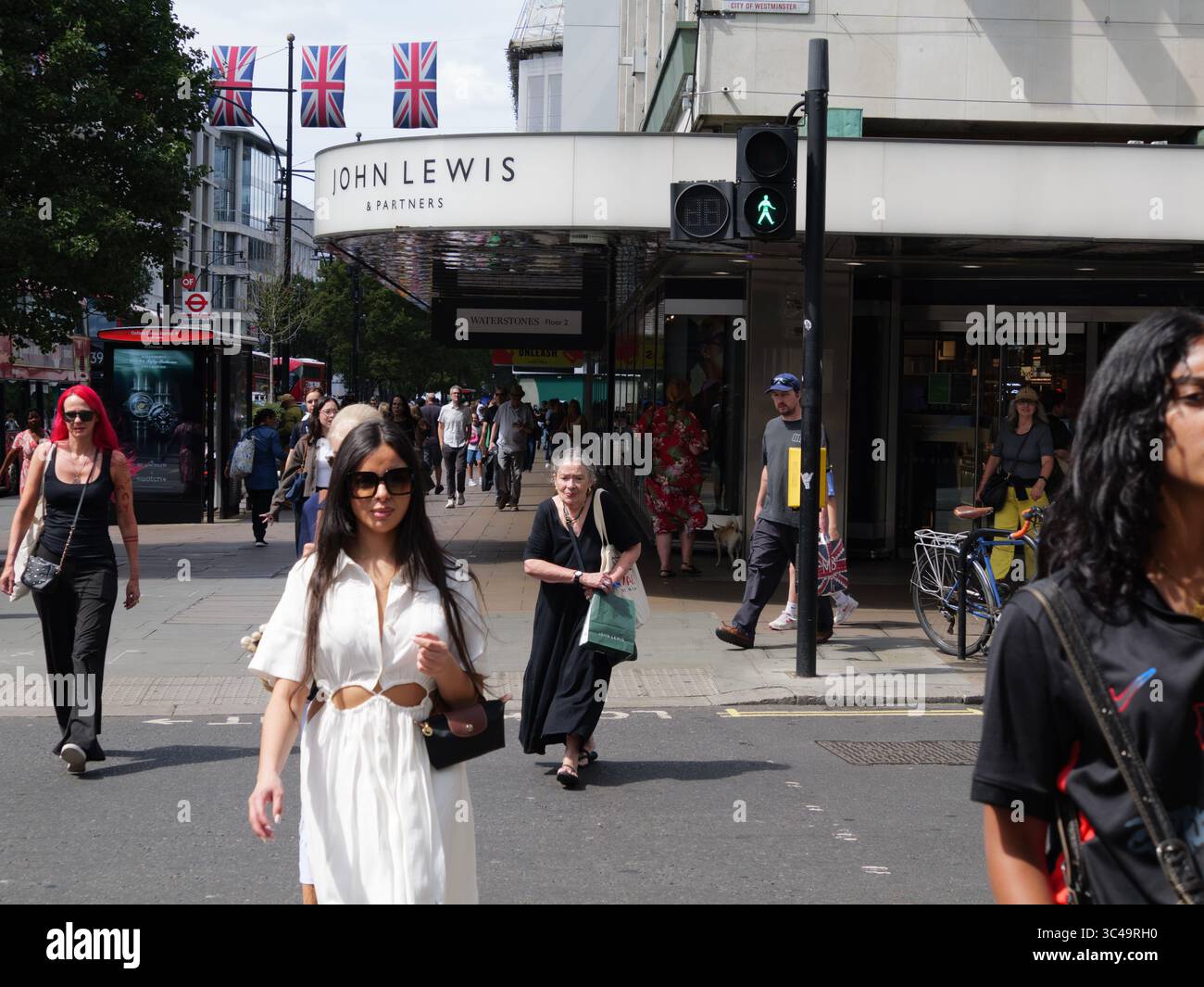 Shoppers walk past the John Lewis department store on Oxford Street, London, UK, with Union Jack flags flying above the bustling high street. Stock Photo