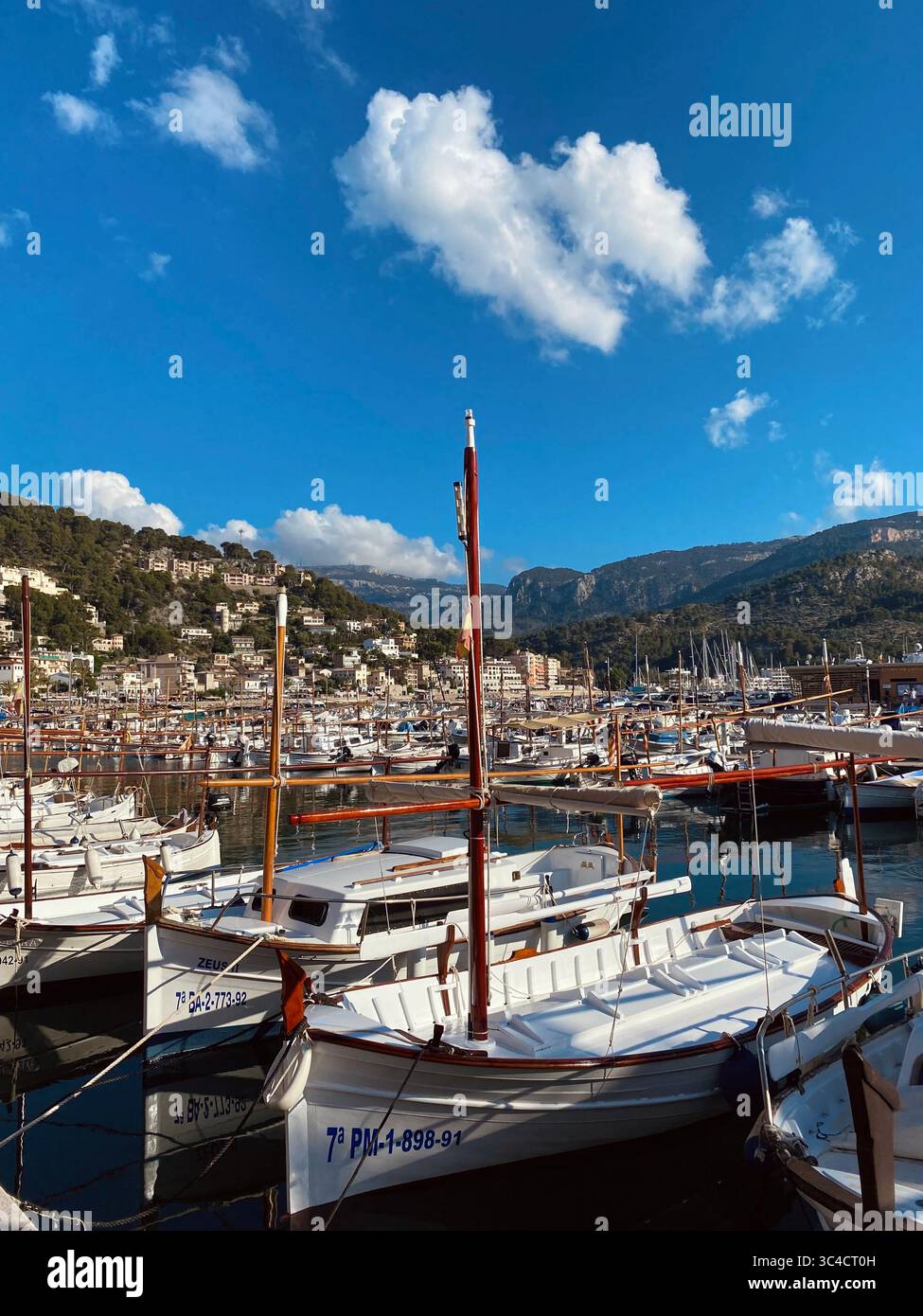 Traditional fishing boats anchored in the marina of Port de Sòller, Mallorca, Spain, with a bright blue sky and scattered small clouds on a summer day - Smartphone Captured Stock Image