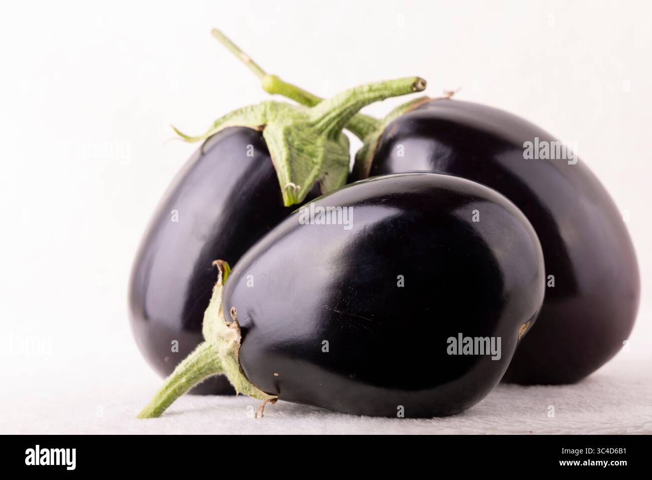 Three black eggplants are sitting on a white surface. The eggplants are large and have a shiny black skin. The stems are green and point upwards. The Stock Photo