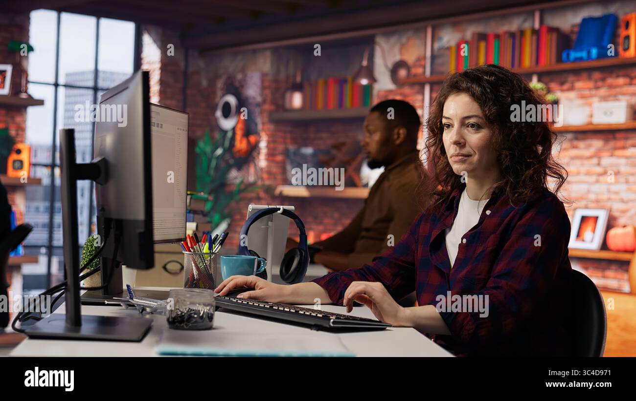 Smiling woman using programming languages on PC in IT startup office to build applications. Upbeat programmer using computer in software development agency to code, camera B Stock Photo