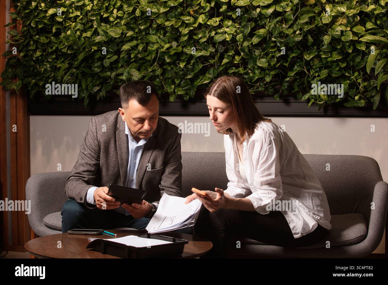 Two business colleagues discuss documents intently on a modern sofa. Professional teamwork, thoughtful expressions. Businessman and assistante . Stock Photo