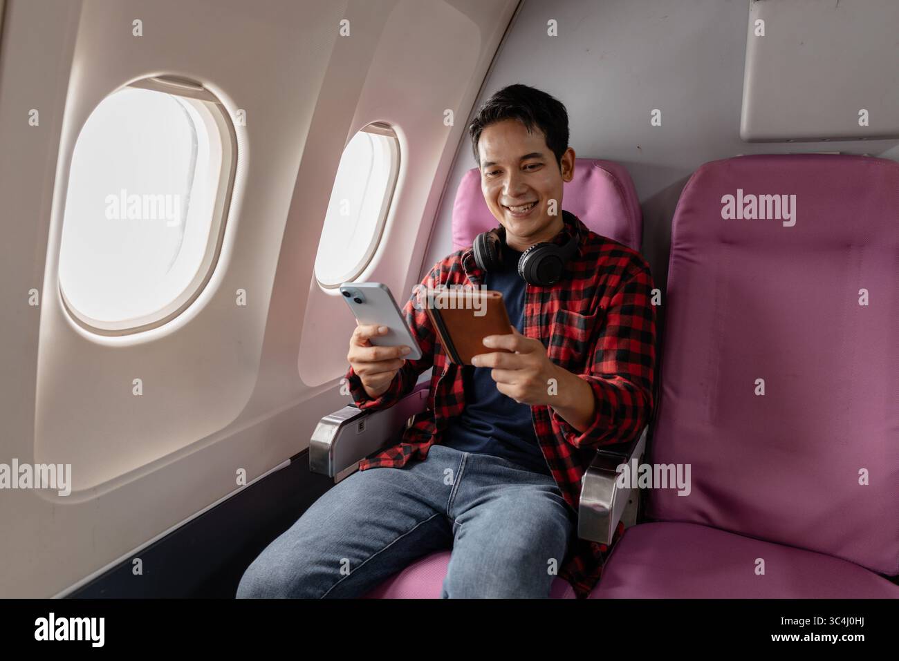 A young man sits on an airplane, enjoying his flight while checking his smartphone and passport. He appears relaxed and content, reflecting the modern Stock Photo