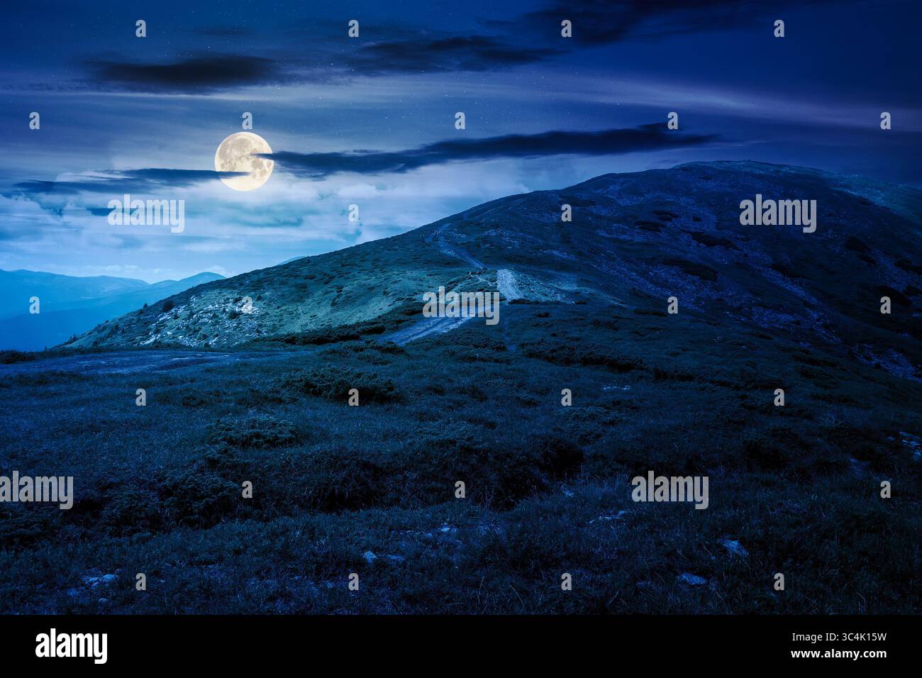 wonderful mountain landscape with rolling hills at night. summer outdoor adventure with path to the top in full moon light. dark blue sky with clouds. Stock Photo