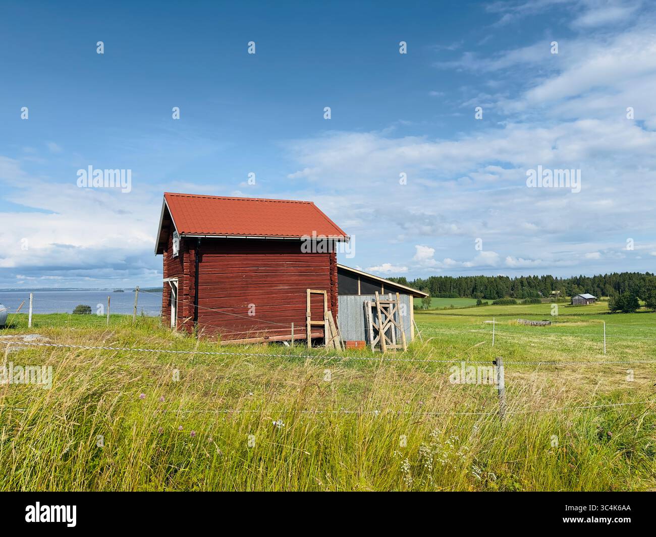 A charming red wooden house stands peacefully in a lush, green field under a blue sky. Stock Photo