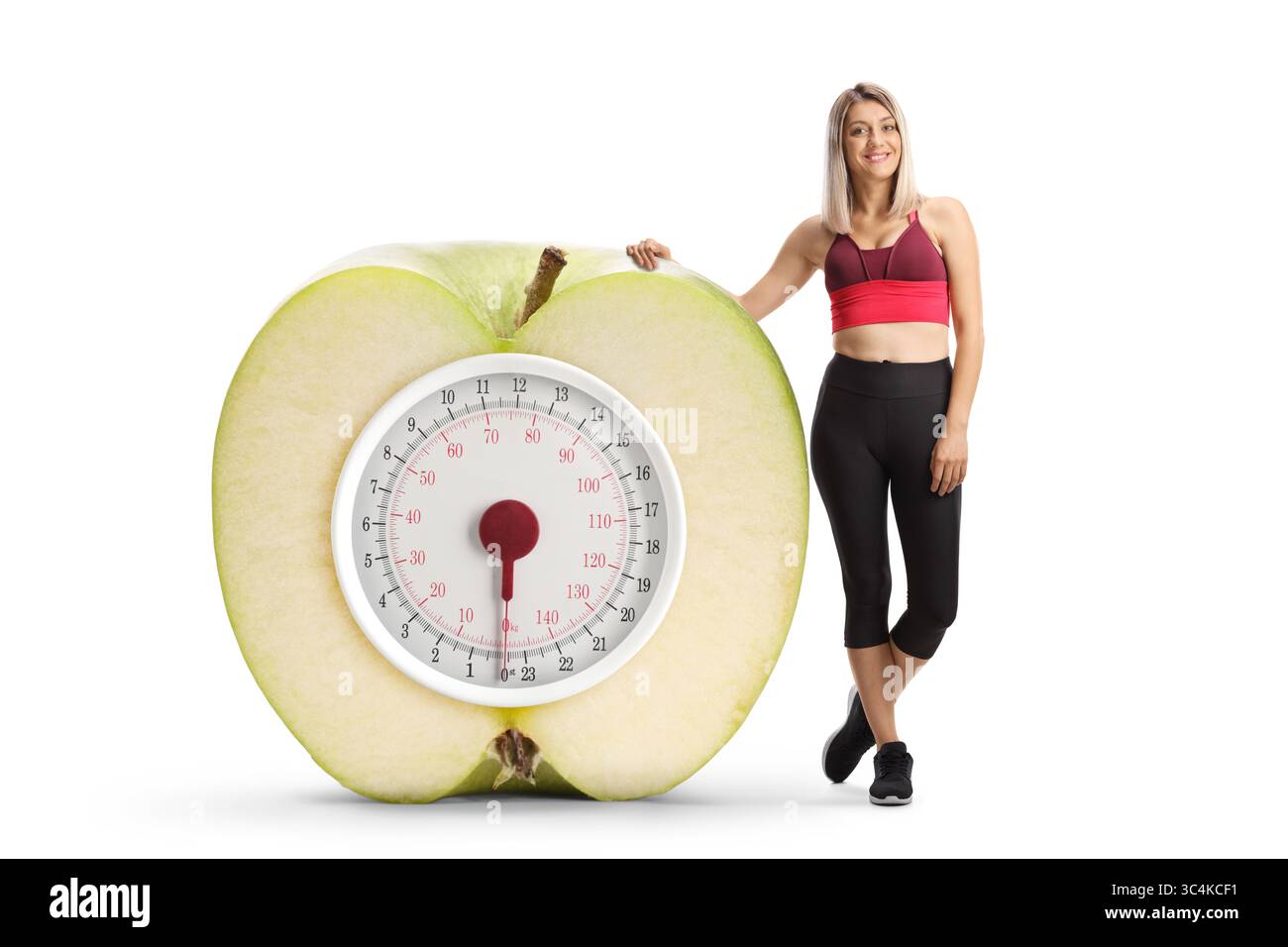 Young woman in sports clothing leaning on a big weight scale inside an apple isolated on white background Stock Photo