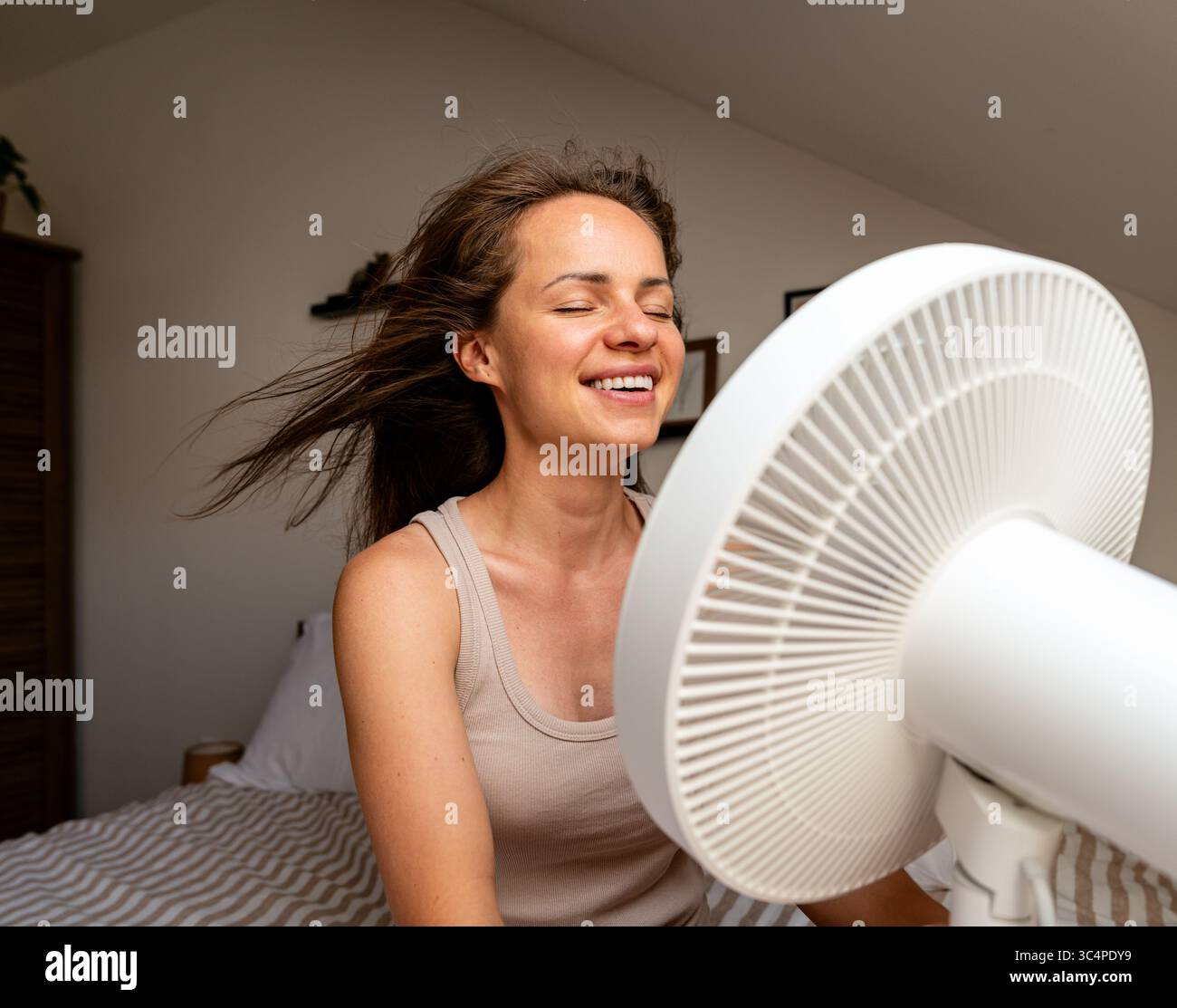 Smiling young woman enjoying cool breeze from electric fan on a hot summer day Stock Photo