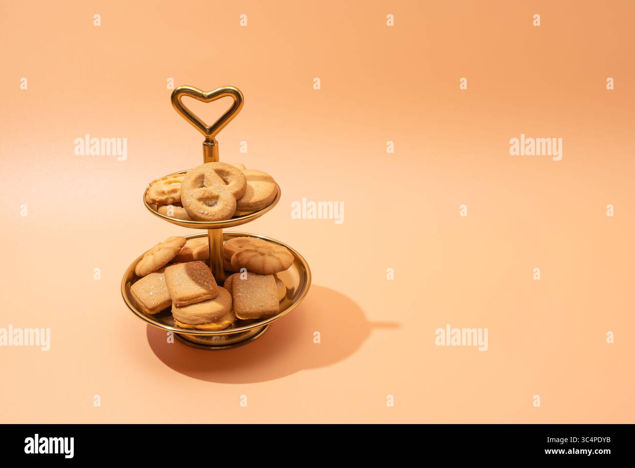 A golden two-tier cookie stand with Danish butter cookies captured from a slight angle. The heart-shaped handle and peach-toned background give a cozy Stock Photo