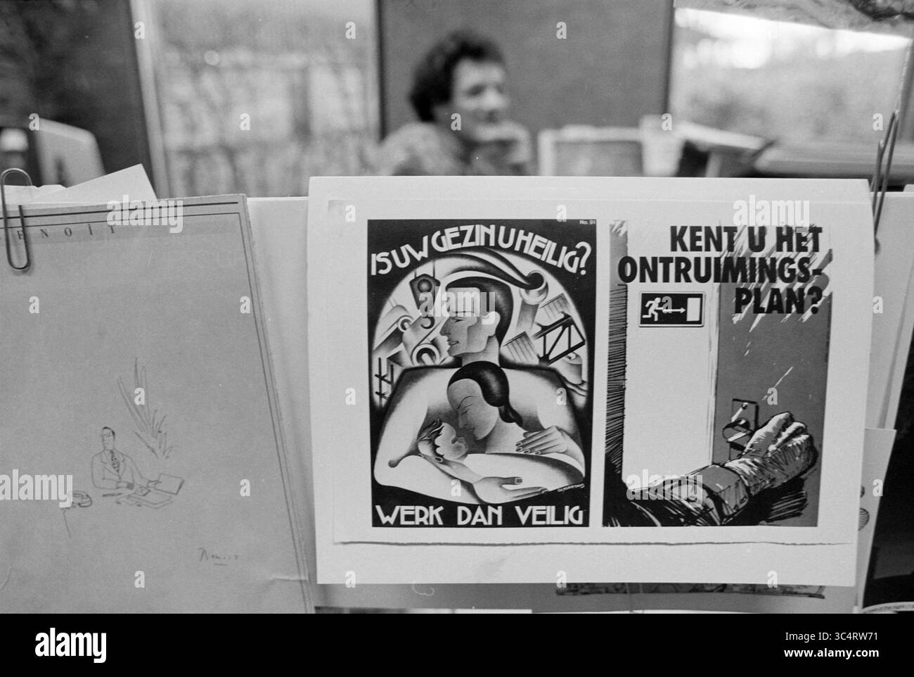 Old educational posters, 00-03-1992 Whizgle News, Dutch Desk, The Netherlands, 1950-2000 A collection of posters and sketches is displayed on a wall, featuring a safety-themed illustration promoting secure work environments, alongside a question about an evacuation plan. In the background, a person is partially visible, engaged in a conversation. Stock Photo