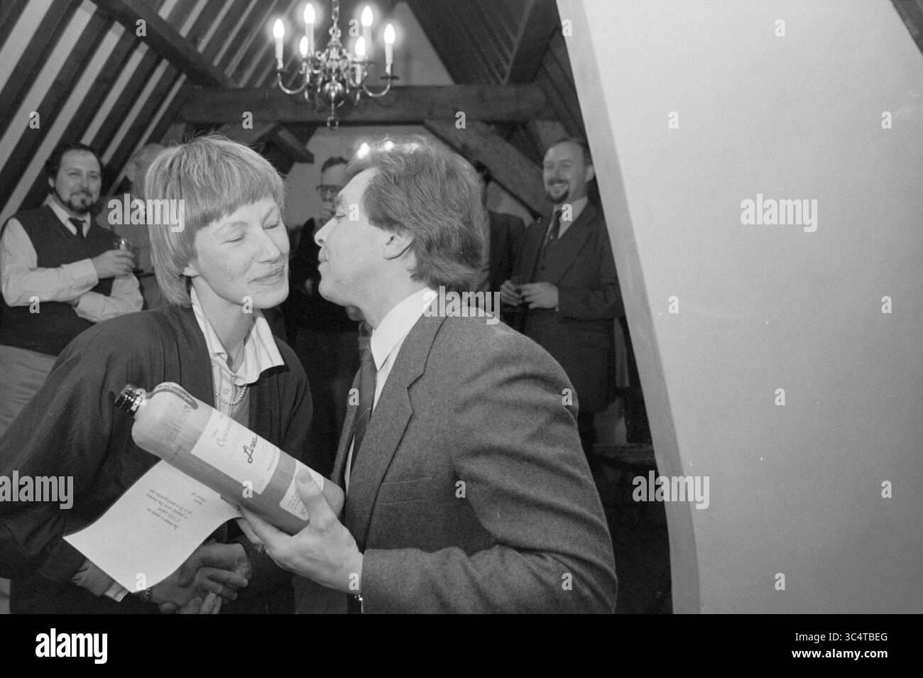 50th member Linea Begijnenhof, Persons, Haarlem, Begijnhof, The Netherlands, 10-02-1987 Whizgle News, Dutch Desk, The Netherlands, 1950-2000 A man and a woman share a playful kiss while holding a bottle of wine, surrounded by cheerful guests enjoying the celebration in a rustic setting. Stock Photo