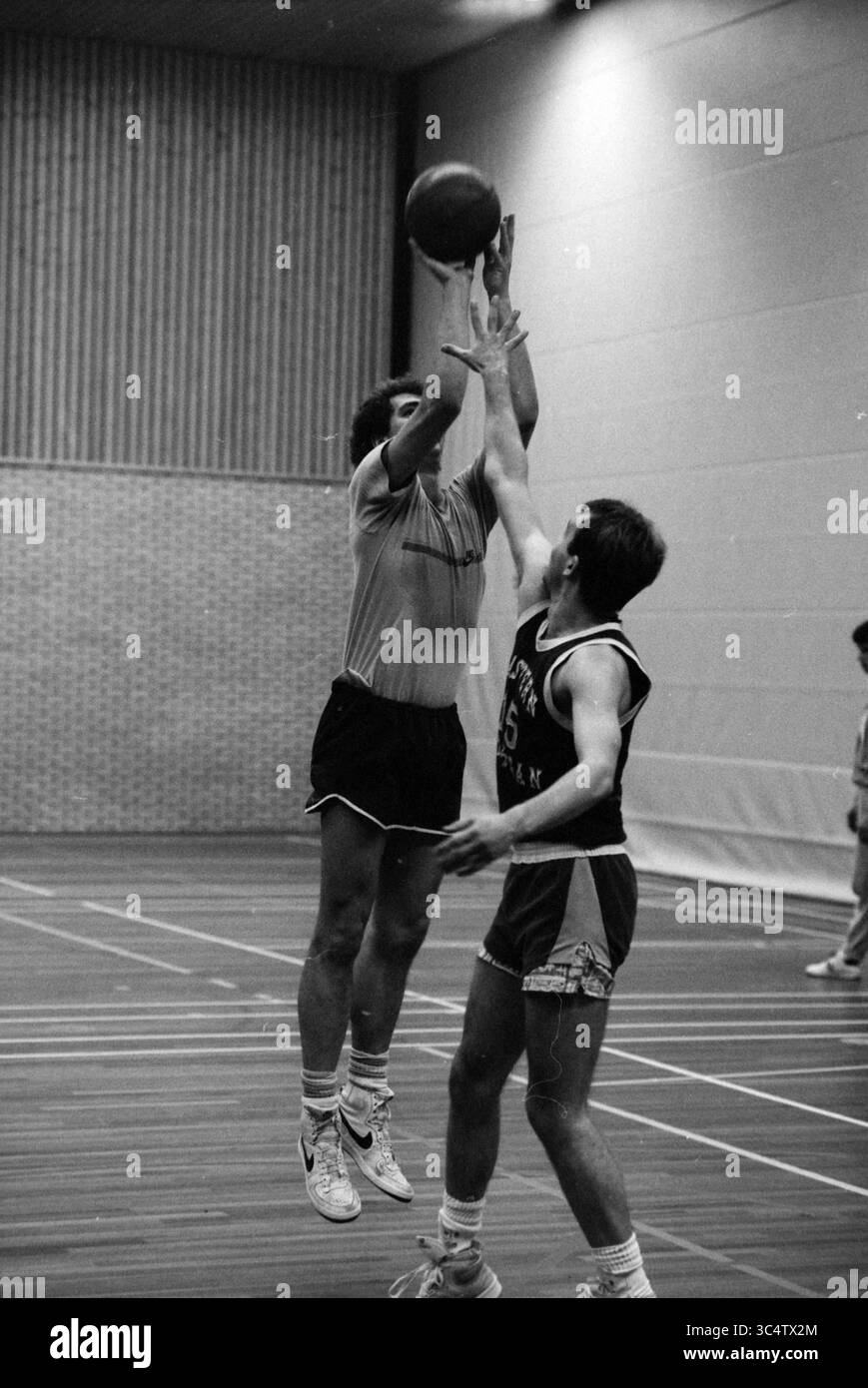 Basketball training Whizgle News, Dutch Desk, The Netherlands, 1950-2000 Two players are engaged in a dynamic basketball game, with one jumping to shoot the ball while the other attempts to block the shot. The indoor court is marked with wooden floors and surrounding players are visible in the background, intensifying the competitive atmosphere. Stock Photo