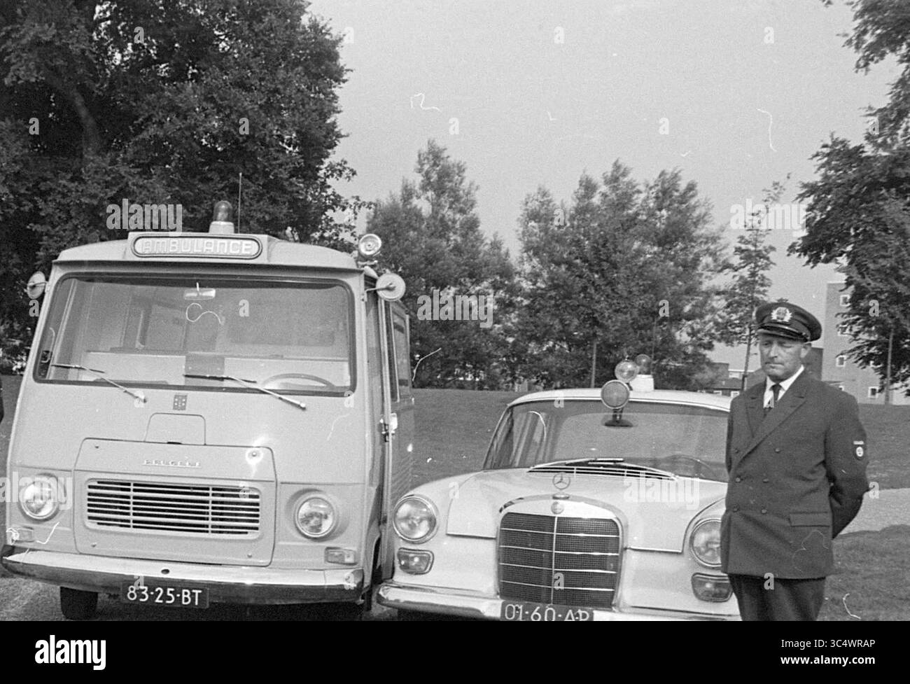 Ambulance with staff, G.G.D., GG and GD, 17-07-1968 Whizgle News, Dutch Desk, The Netherlands, 1950-2000 A vintage ambulance and a classic police car are parked side by side, while a uniformed officer stands nearby, surveying the scene in a serene outdoor setting surrounded by trees. Stock Photo