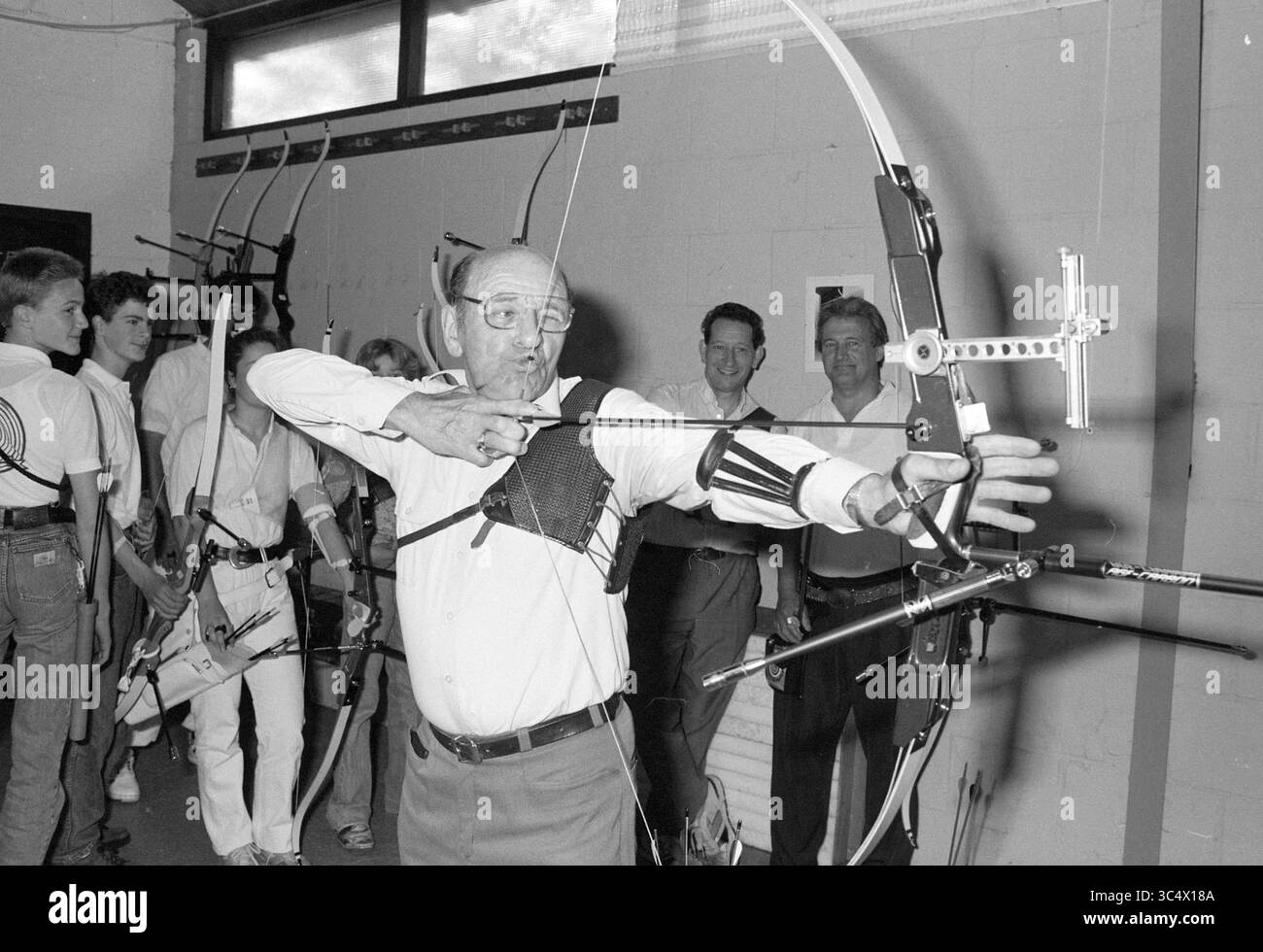 Jan Koens 50 years as an archer, 22-06-1991 Whizgle News, Dutch Desk, The Netherlands, 1950-2000 An elderly archer demonstrates his skills with a bow, focusing intently as he prepares to shoot. Behind him, a group of younger archers observe, showcasing their own bows and equipment in a lively indoor setting. Stock Photo