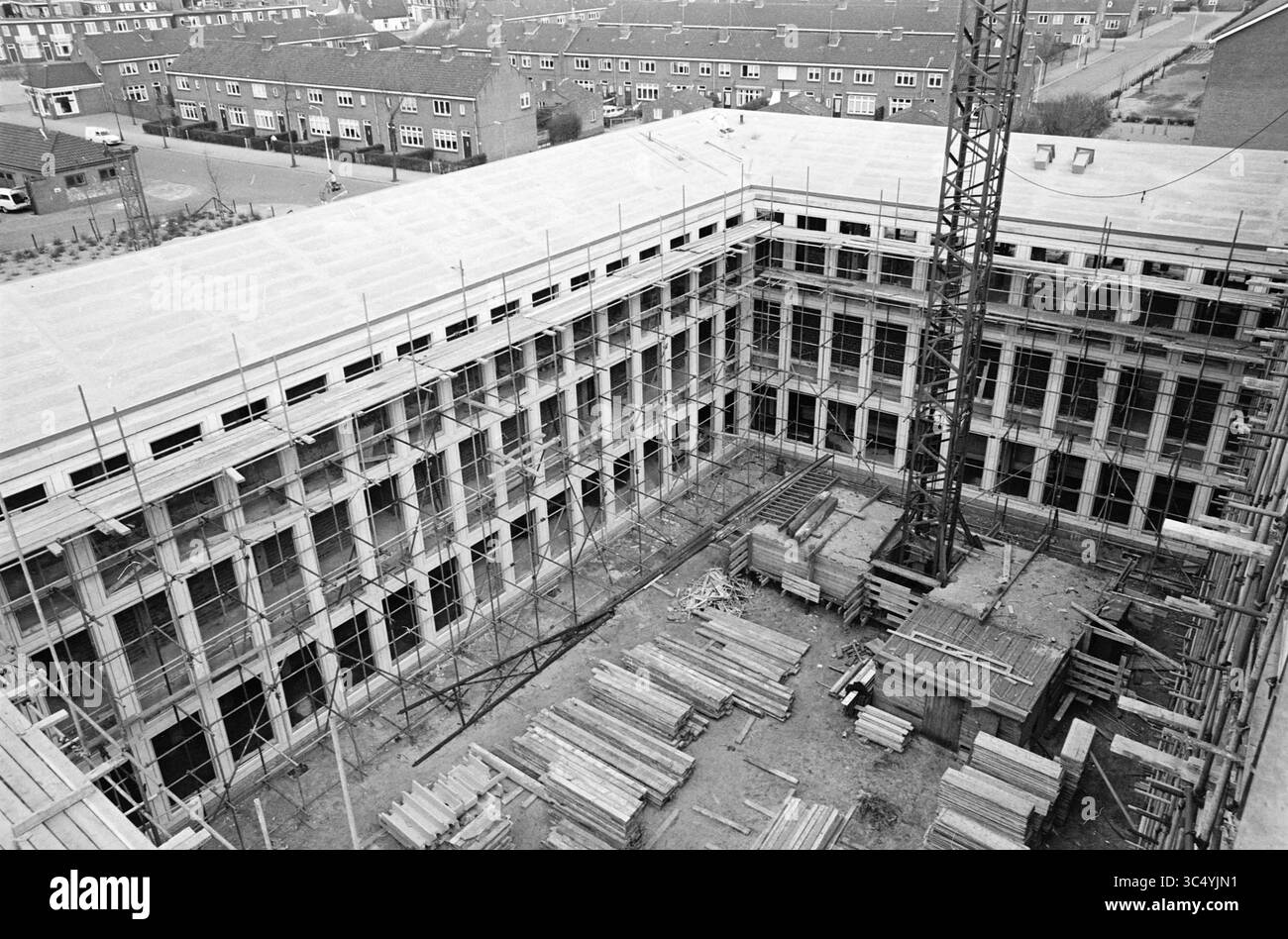 Beautiful photos in the areas of IJmuiden, IJmuiden, Velsen, 20-04-1964 Whizgle News, Dutch Desk, The Netherlands, 1950-2000 A large construction site showcases a building frame under development, featuring scaffolding, stacks of wooden materials, and a towering crane. Surrounding the site are several residential buildings, indicating an urban environment. Stock Photo