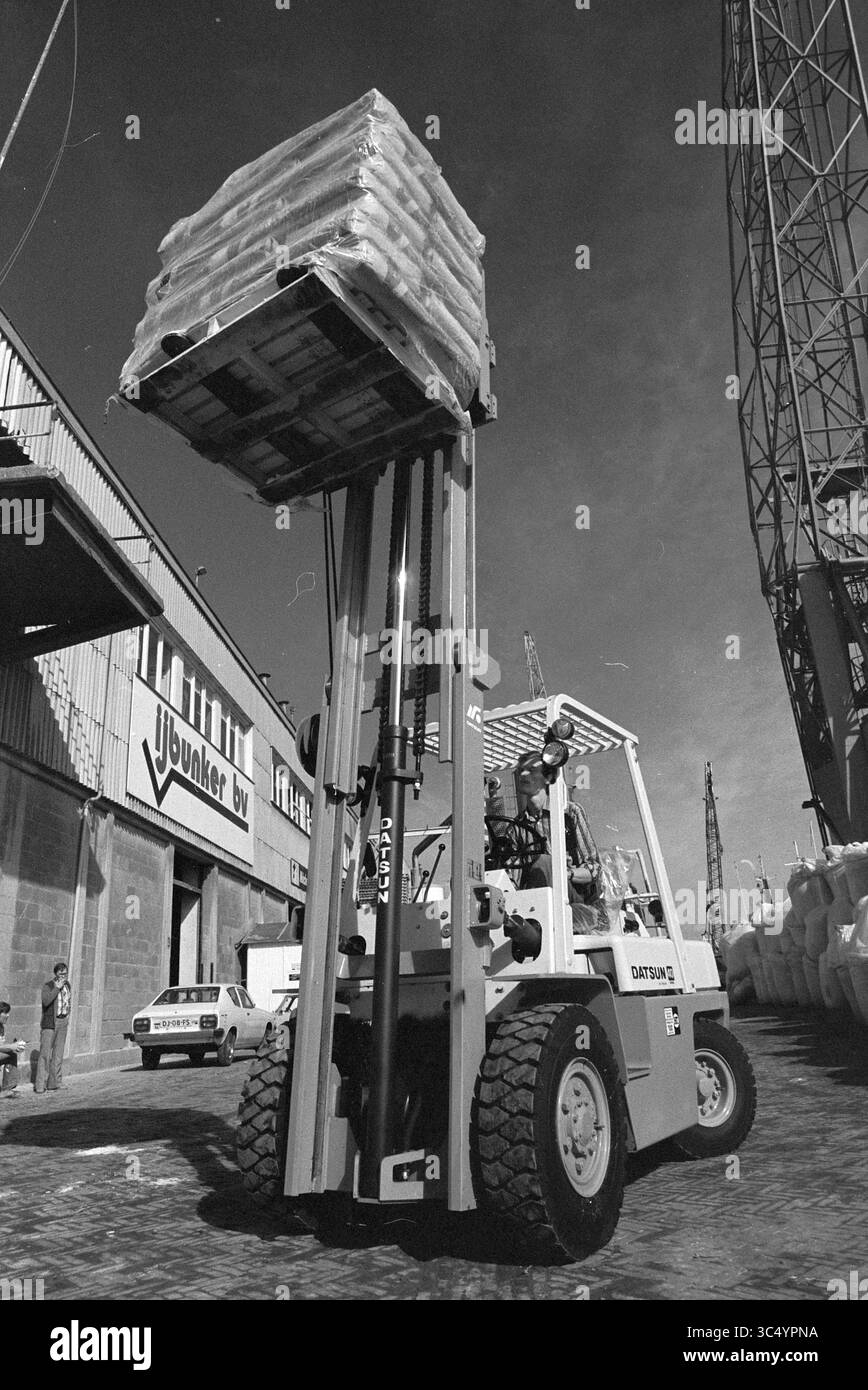 Turbo front forklift IJ-Bunker, Work, 13-08-1980 Whizgle News, Dutch Desk, The Netherlands, 1950-2000 A forklift lifts a high stack of pallets loaded with bags, positioned near industrial buildings and a large tower. The scene captures an active work environment, with vehicles parked in the background and machinery visible. Stock Photo