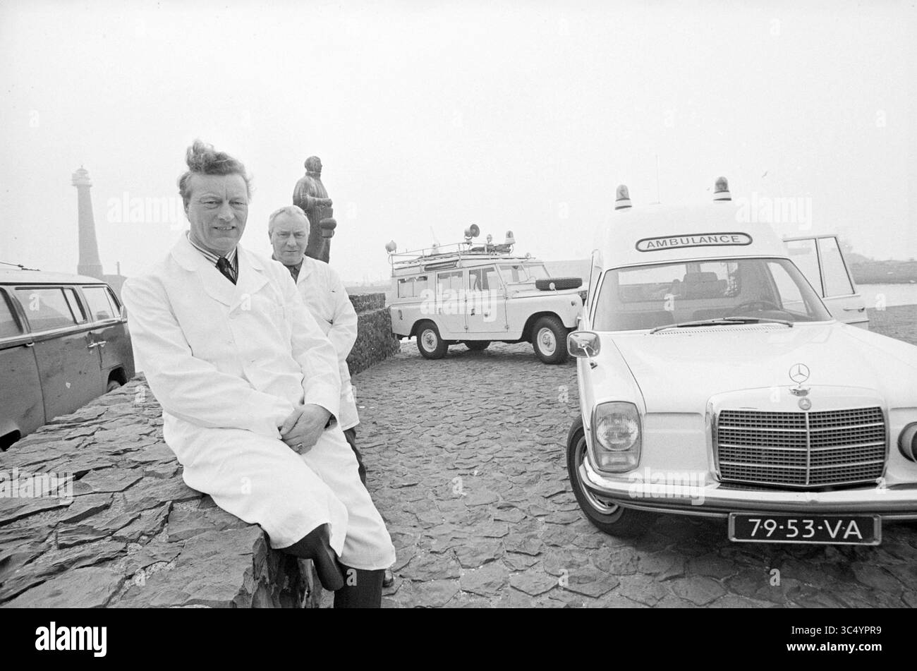Ambulance personnel at their car., IJmuiden, Sluisplein, The Netherlands Whizgle News, Dutch Desk, The Netherlands, 1950-2000 Two men in white lab coats stand beside a vintage ambulance parked on a stone wall, with a lighthouse visible in the background. One man sits casually on the wall, while the other stands nearby, both appearing engaged in conversation or observation. The scene conveys a sense of calm and professional demeanor amidst a coastal setting. Stock Photo