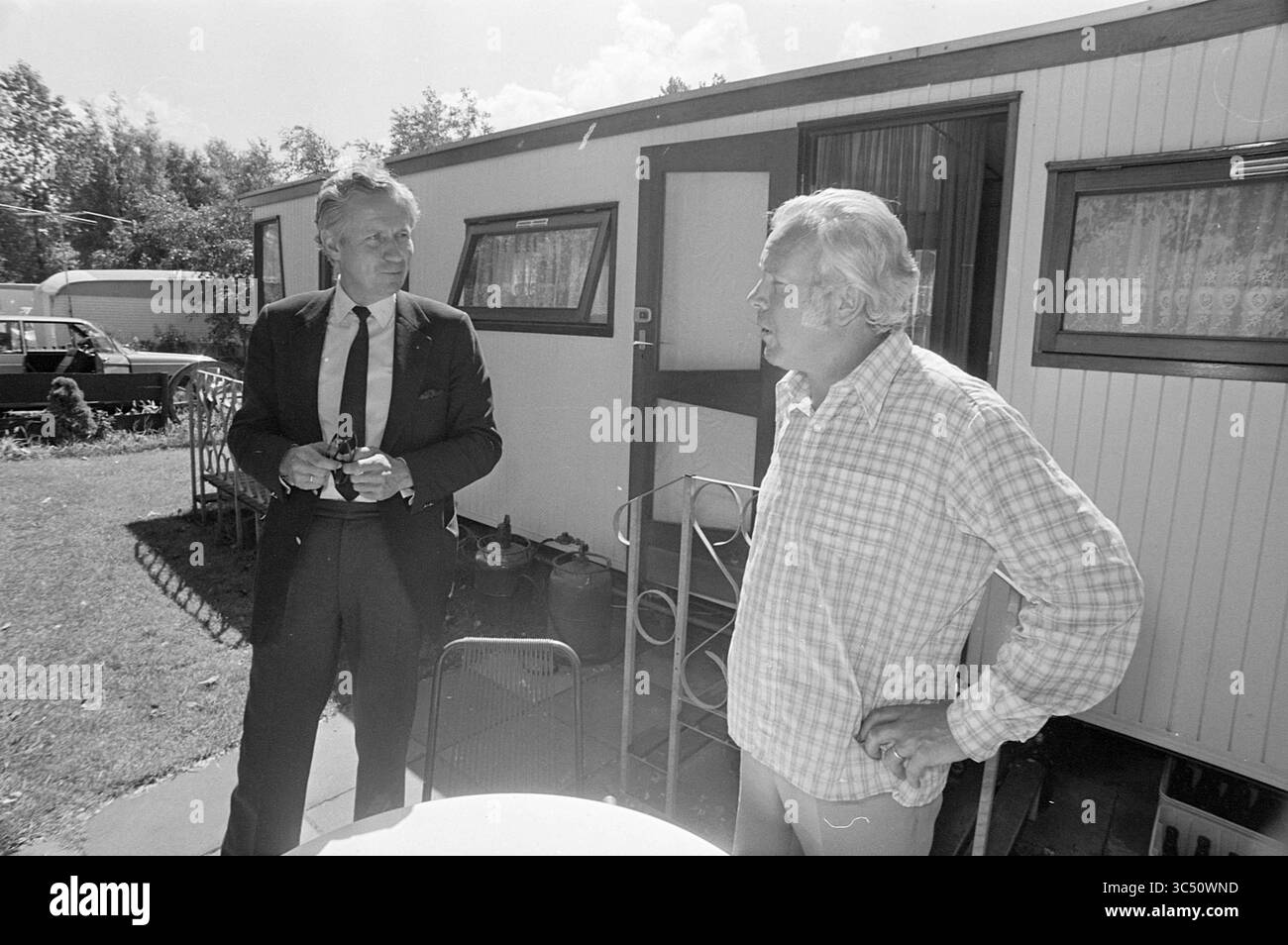 Duinrell camping in Wassenaar, Kampen, campsites, camp site, camping, camping hut, Tourism, tourists, 26-07-1974 Whizgle News, Dutch Desk, The Netherlands, 1950-2000 Two men engage in conversation outside a mobile home, with one dressed in a suit and the other in a casual plaid shirt. The setting features grass and a few lawn chairs, suggesting a relaxed outdoor environment. Trees and additional vehicles are visible in the background. Stock Photo