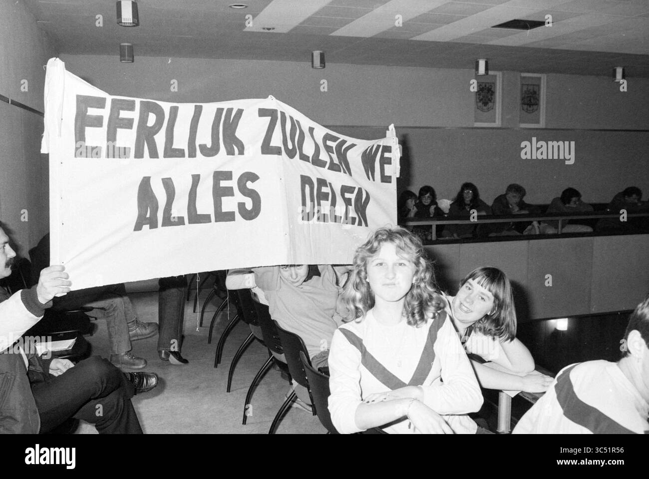 Velsen council chamber, activists in the public gallery with a banner, IJmuiden, The Netherlands, 20-12-1979 Whizgle News, Dutch Desk, The Netherlands, 1950-2000 A group of individuals gathered in a public space, with one person prominently holding a large banner that reads 'Eerlijk zullen we alles delen,' while others sit nearby, some smiling and engaging with the scene. The atmosphere suggests a moment of protest or expression of solidarity. Stock Photo
