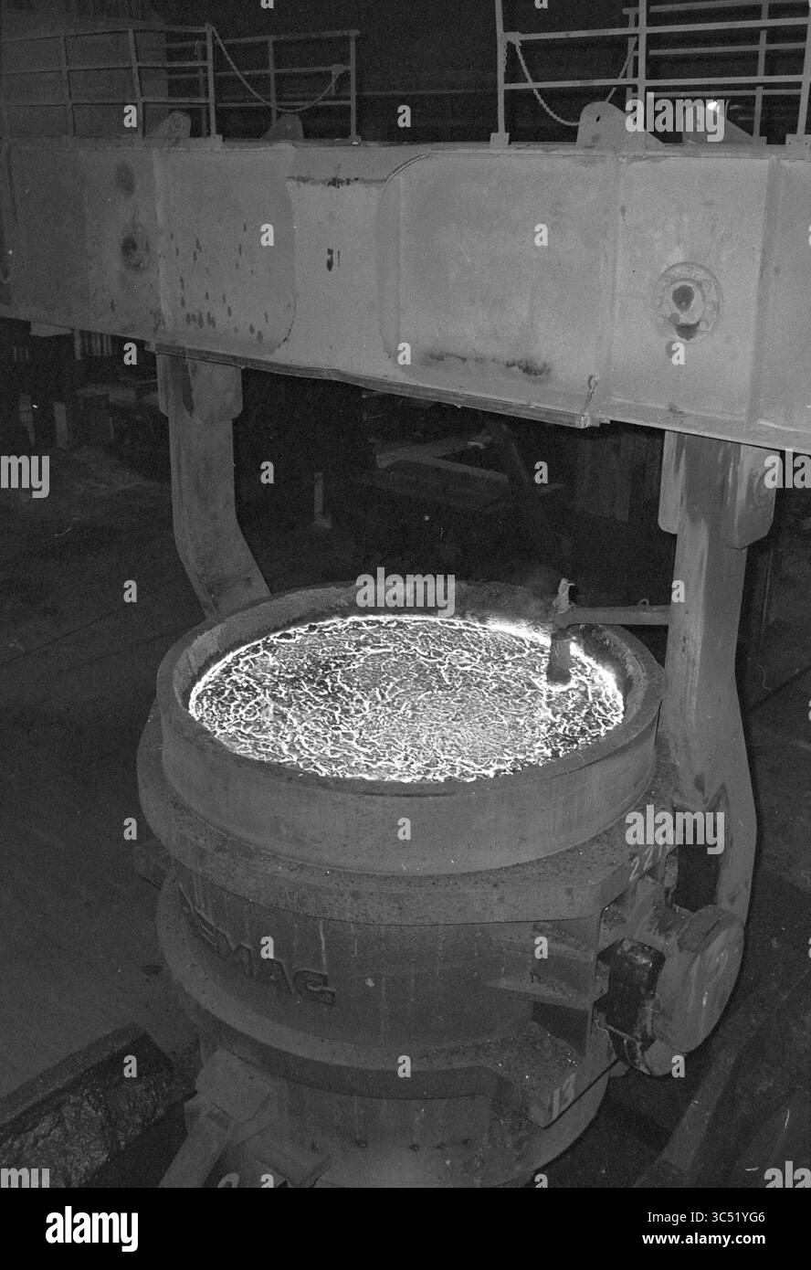 Reduction of working hours Hoogovens + company photos, Hoogovens, 12-11-1980 Whizgle News, Dutch Desk, The Netherlands, 1950-2000 A large, heavy-duty vessel filled with molten metal glows brightly, surrounded by industrial machinery that towers above it. The surface of the molten metal exhibits a textured pattern, reflecting the intense heat and energy of the foundry environment. Stock Photo