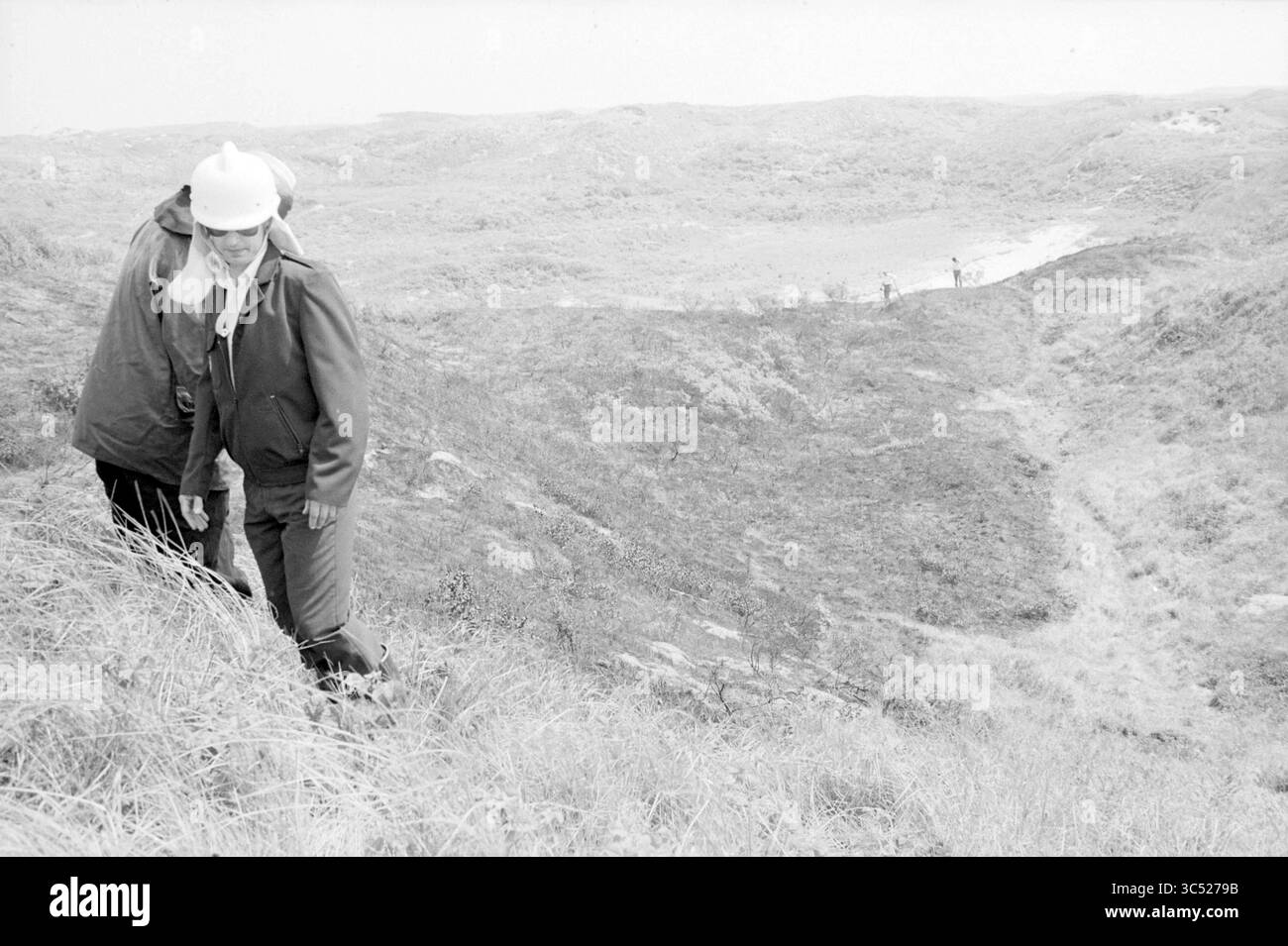 Dune fire Parnassia Bloemendaal, Fires, fire brigade, Overveen, Parnassiaweg, 19-06-1986 Whizgle News, Dutch Desk, The Netherlands, 1950-2000 Two individuals carefully navigate through a hilly landscape, observing the terrain around them. In the distance, additional figures can be seen exploring the area, while the natural surroundings are characterized by rolling hills and sparse vegetation. Stock Photo