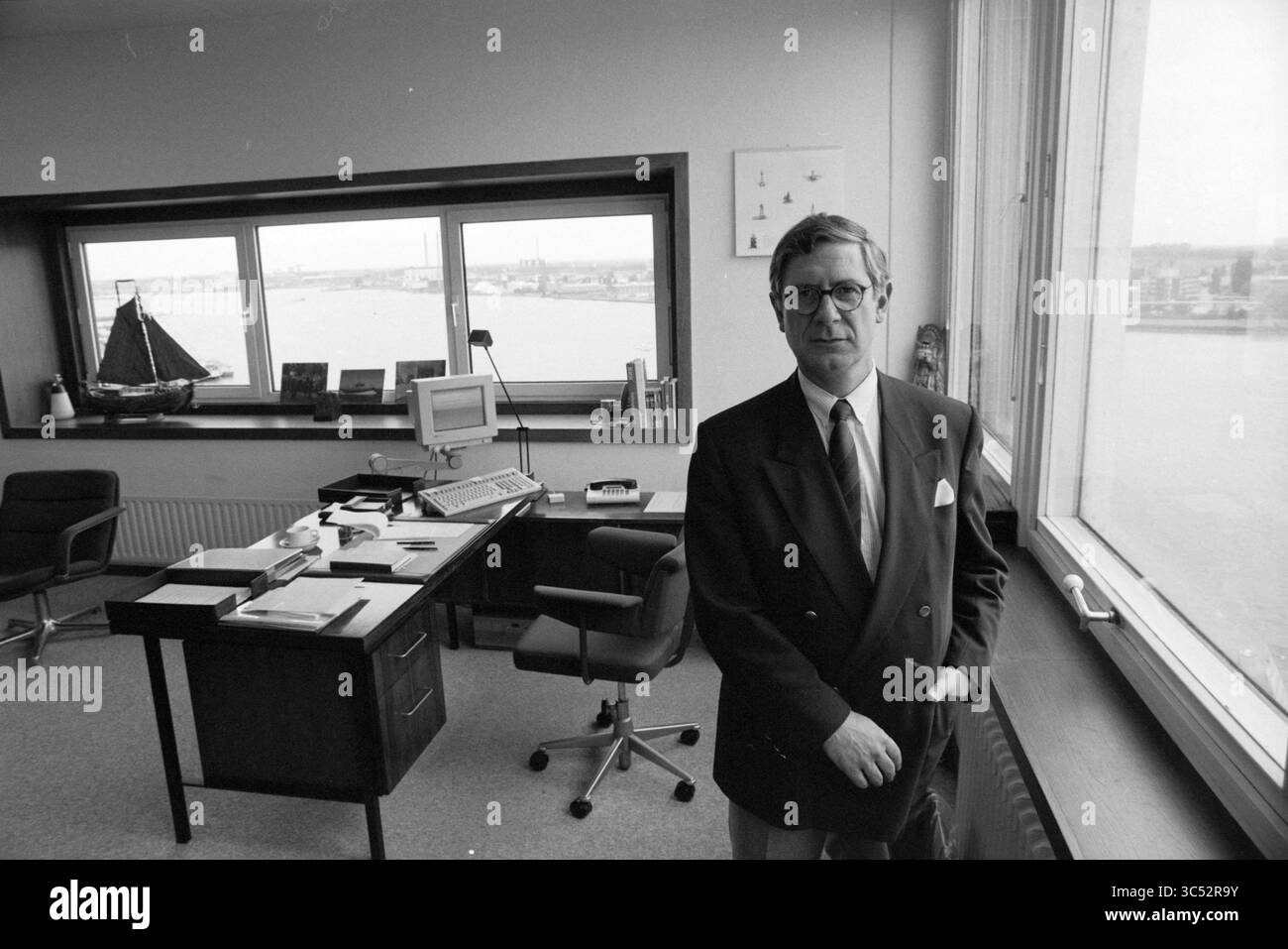 Director of Hennel Port Authority, Port Building, De Ruijterkade, Amsterdam, Amsterdam, De Ruijterkade, The Netherlands, 25-07-1990 Whizgle News, Dutch Desk, The Netherlands, 1950-2000 A businessman stands confidently in a well-organized office, with a large window revealing a view of the water and city beyond. The space features a desk with a computer, stationery, and a model ship, reflecting a professional atmosphere. Stock Photo