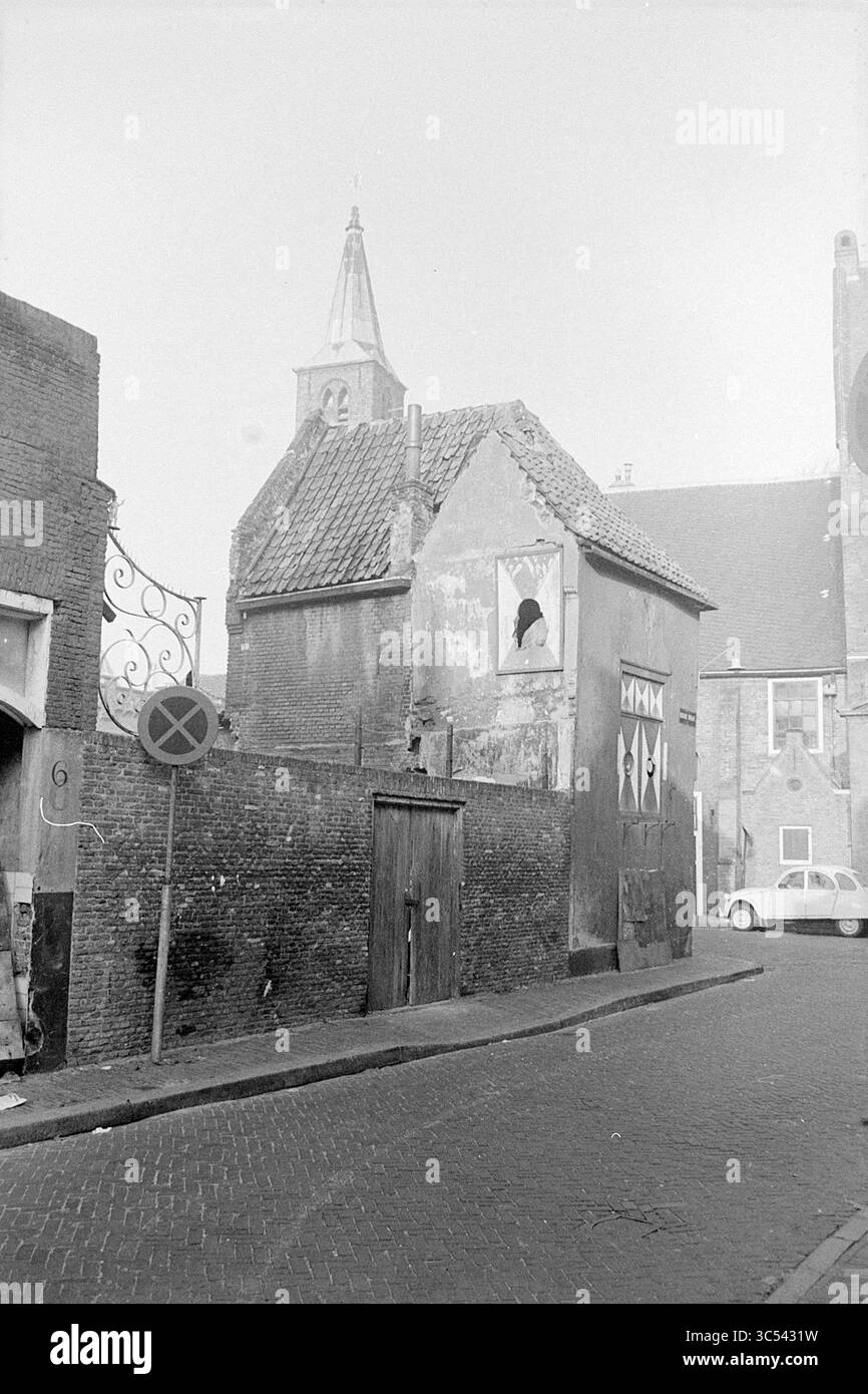 Ext. Begijnehof 20, Exterior, Haarlem, Begijnhof, The Netherlands, 11-12-1969 Whizgle News, Dutch Desk, The Netherlands, 1950-2000 A narrow street lined with quaint buildings, one featuring a broken window and a distinctive steeple. An old car is parked nearby, adding to the scene's nostalgic charm. Stock Photo