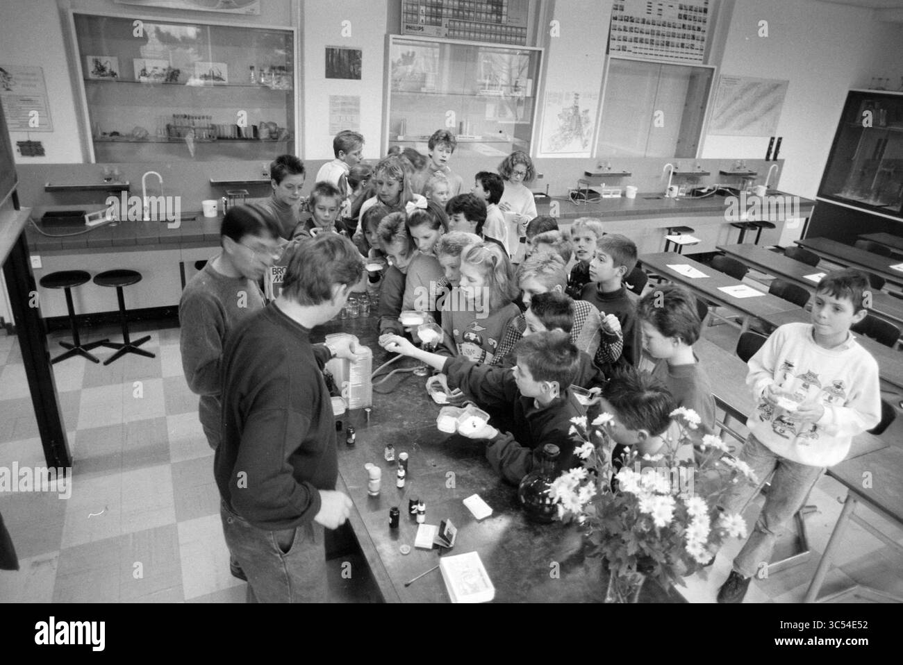 Information days RK Leeuwenhoek School Noordwijkerhout, Schools, Noordwijkerhout, 25-01-1989 Whizgle News, Dutch Desk, The Netherlands, 1950-2000 A group of excited children huddle around a table in a classroom, eagerly watching an adult demonstrate a science experiment, with various materials and a flower arrangement nearby. Stock Photo