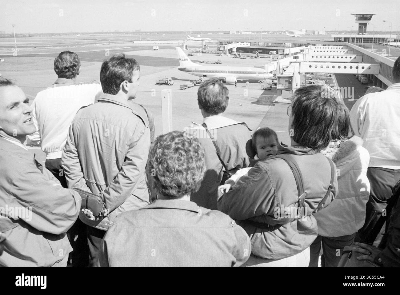 Viewers at Schiphol, Schiphol, 00-04-1990 Whizgle News, Dutch Desk, The Netherlands, 1950-2000 A group of people gathers by a large window at an airport, observing the bustling tarmac filled with parked airplanes and ground crew preparing for departures. Some individuals are engaging in conversation, while others gaze intently outside, capturing a moment of anticipation and excitement. Stock Photo