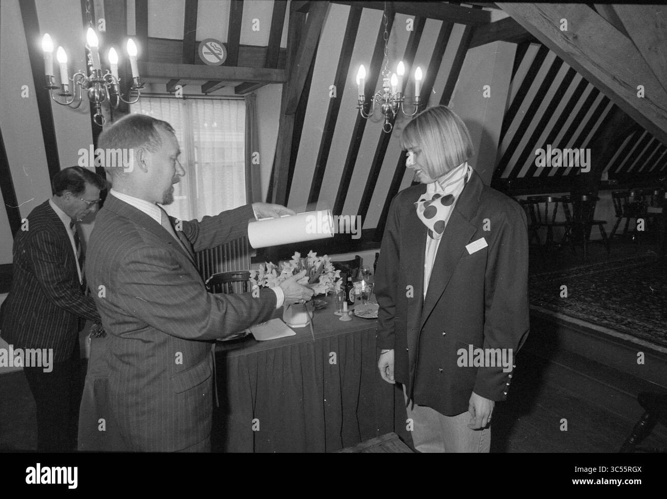 Certificate for the 100th member of the Soos Begijnhof, Haarlem, Begijnhof, The Netherlands, 13-03-1990 Whizgle News, Dutch Desk, The Netherlands, 1950-2000 A man in a suit presents a rolled document to a woman wearing a stylish outfit with a scarf, while another individual looks on in a room characterized by wooden beams and elegant decor. Stock Photo