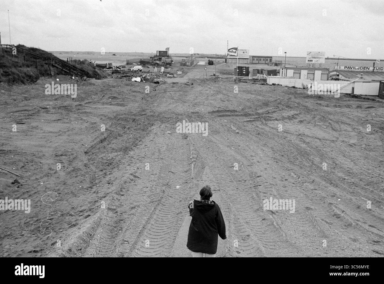 Remains of the North Sea beach pavilion, 00-00-1993 Whizgle News, Dutch Desk, The Netherlands, 1950-2000 A person walks along a sandy expanse that stretches into an empty landscape, marked by tire tracks and scattered debris, with distant signs indicating a once-bustling area. The sky is overcast, adding to the scene's desolate atmosphere. Stock Photo