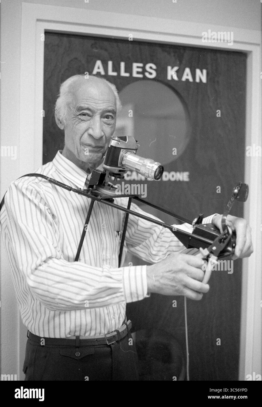 Mr. Gatsonides (speedometer inventor), Portraits, 31-10-1990 Whizgle News, Dutch Desk, The Netherlands, 1950-2000 An older man stands confidently with a camera mounted on a contraption designed for steady shots, showcasing a blend of creativity and engineering. The door behind him features the phrase 'ALLES KAN,' suggesting a theme of possibility or capability. Stock Photo