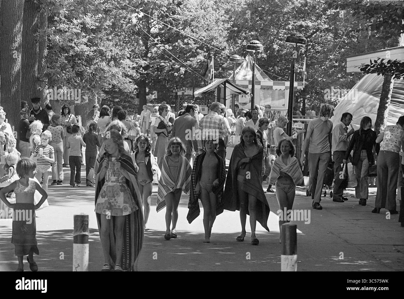 Duinrell camping in Wassenaar, Kampen, campsites, camp site, camping, camping hut, Tourism, tourists, 26-07-1974 Whizgle News, Dutch Desk, The Netherlands, 1950-2000 A lively scene in a park bustling with families and children enjoying a day out, some wearing colorful outfits while others are wrapped in blankets. In the background, a carousel and various attractions create a festive atmosphere as people stroll along the pathway. Stock Photo