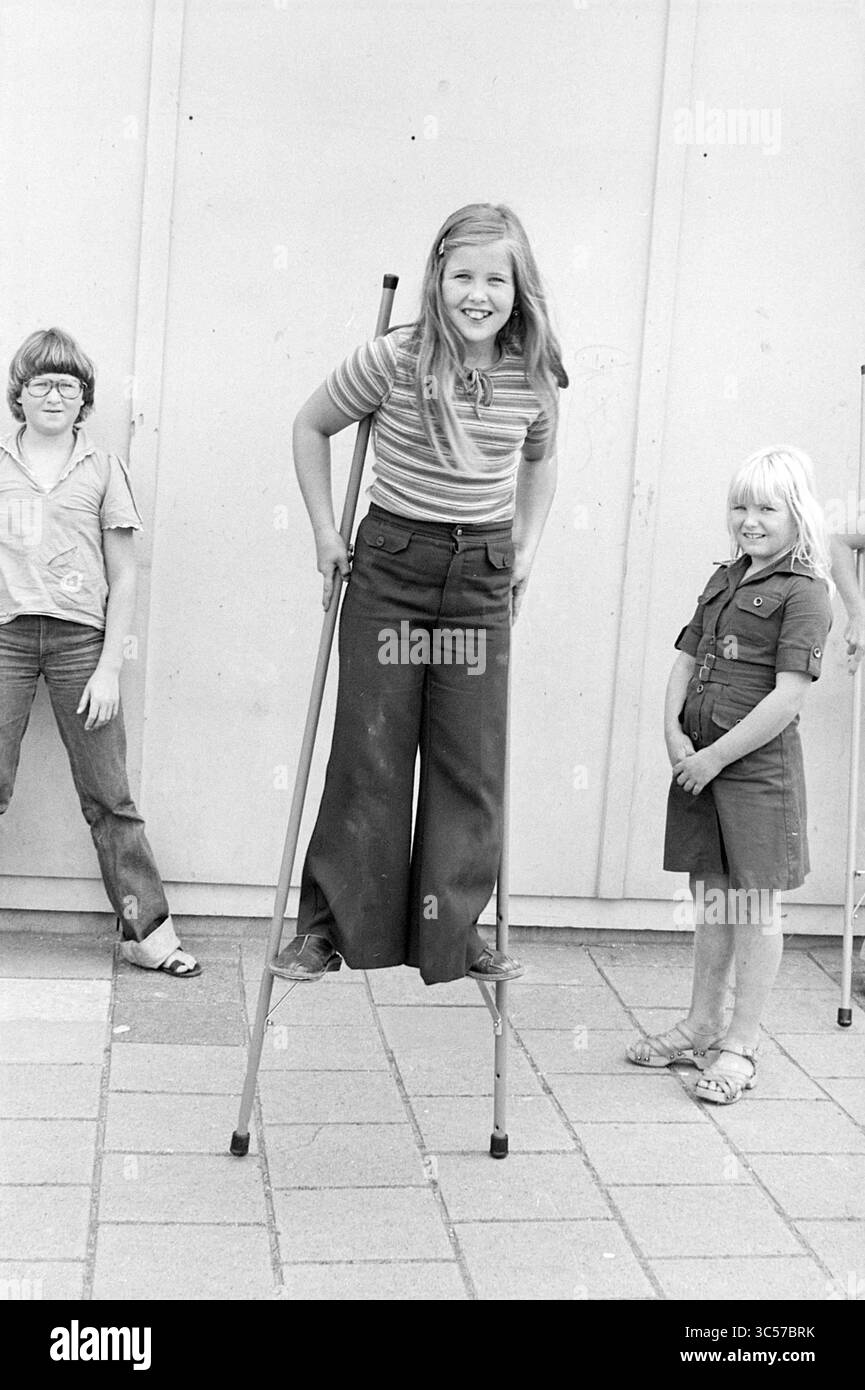 Children's games, Beverwijk, Children, kid, children's homes, children's party, children's, 01-08-1977 Whizgle News, Dutch Desk, The Netherlands, 1950-2000 A cheerful girl stands confidently on stilts, sporting a striped shirt and wide-legged pants. Two friends pose nearby, one wearing glasses and a casual shirt, and the other in a playful jumpsuit, all sharing smiles in a lively outdoor setting. Stock Photo
