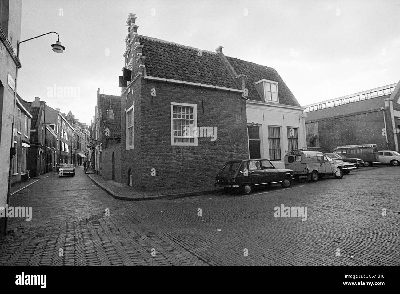 Begijnhof building, Exterior, Haarlem, Begijnhof, The Netherlands, 15-09-1972 Whizgle News, Dutch Desk, The Netherlands, 1950-2000 A quiet street corner features a distinctive brick building with a sloped roof, flanked by parked vehicles. The cobblestone road leads into a charming neighborhood with historical architecture, hinting at the area's character and history. Stock Photo