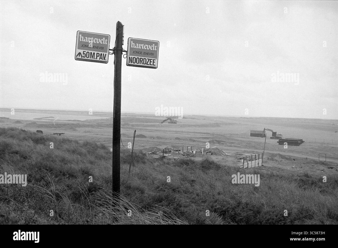 Remains of the North Sea beach pavilion, 00-00-1993 Whizgle News, Dutch Desk, The Netherlands, 1950-2000 A weathered signpost marks the boundary of Hartevelt, offering directional guidance amidst a vast, industrial landscape. In the background, heavy machinery works against a cloudy sky, while mounds of earth and debris hint at ongoing development in the area. Stock Photo