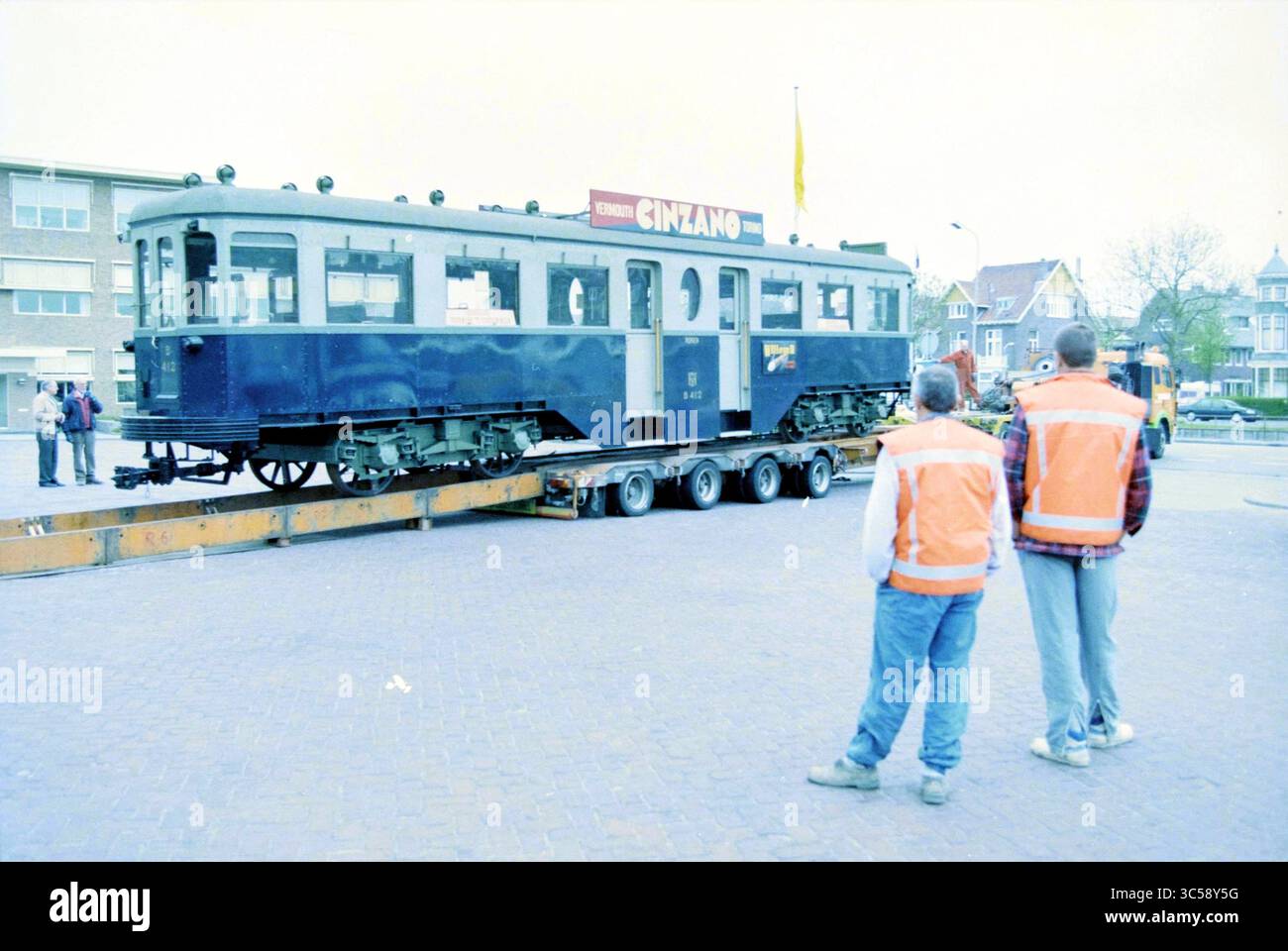 Relocation of the Blue Tram to the new museum of the NZH, Haarlem, Leidsevaart, The Netherlands, 02-05-1996 Whizgle News, Dutch Desk, The Netherlands, 1950-2000 A vintage train carriage is being transported on a flatbed trailer, with two individuals in safety vests observing the scene. Nearby buildings and a flag add context to the bustling environment. Stock Photo
