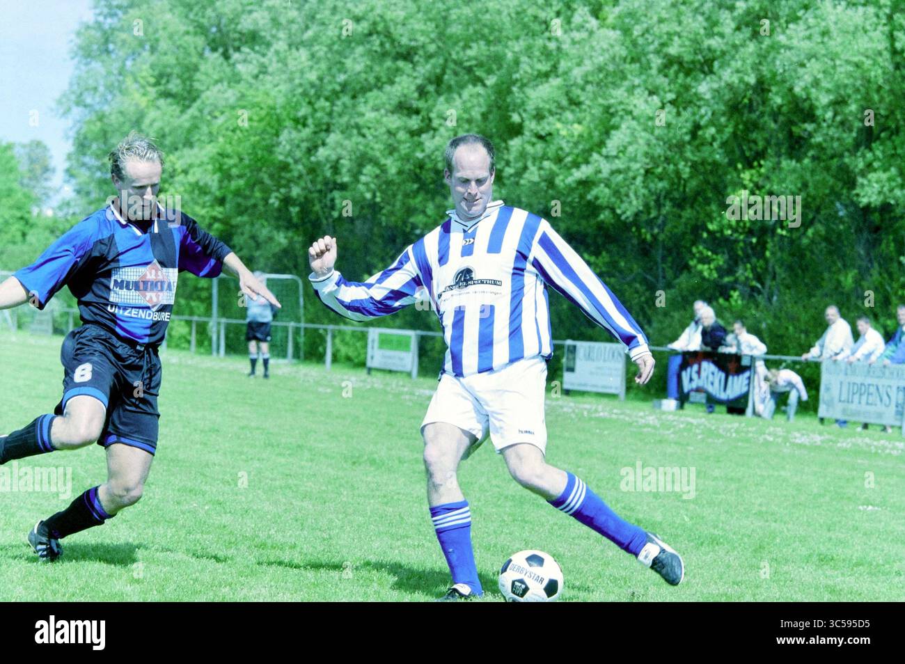 Football: Hillegom - VVZ, Hillegom, 10-05-2003 Whizgle News, Dutch Desk, The Netherlands, 1950-2000 Two soccer players compete for the ball on a grassy field. One player, wearing a blue and white striped jersey, skillfully maneuvers to maintain possession, while the other, in a black kit, attempts to challenge him for the ball. In the background, spectators can be seen cheering on the teams. Stock Photo