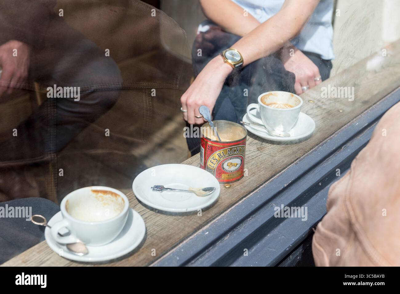 Candid view through cafe window with coffee cups, saucers, and hand stirring vintage sugar tin, capturing relaxed coffee culture in London Stock Photo