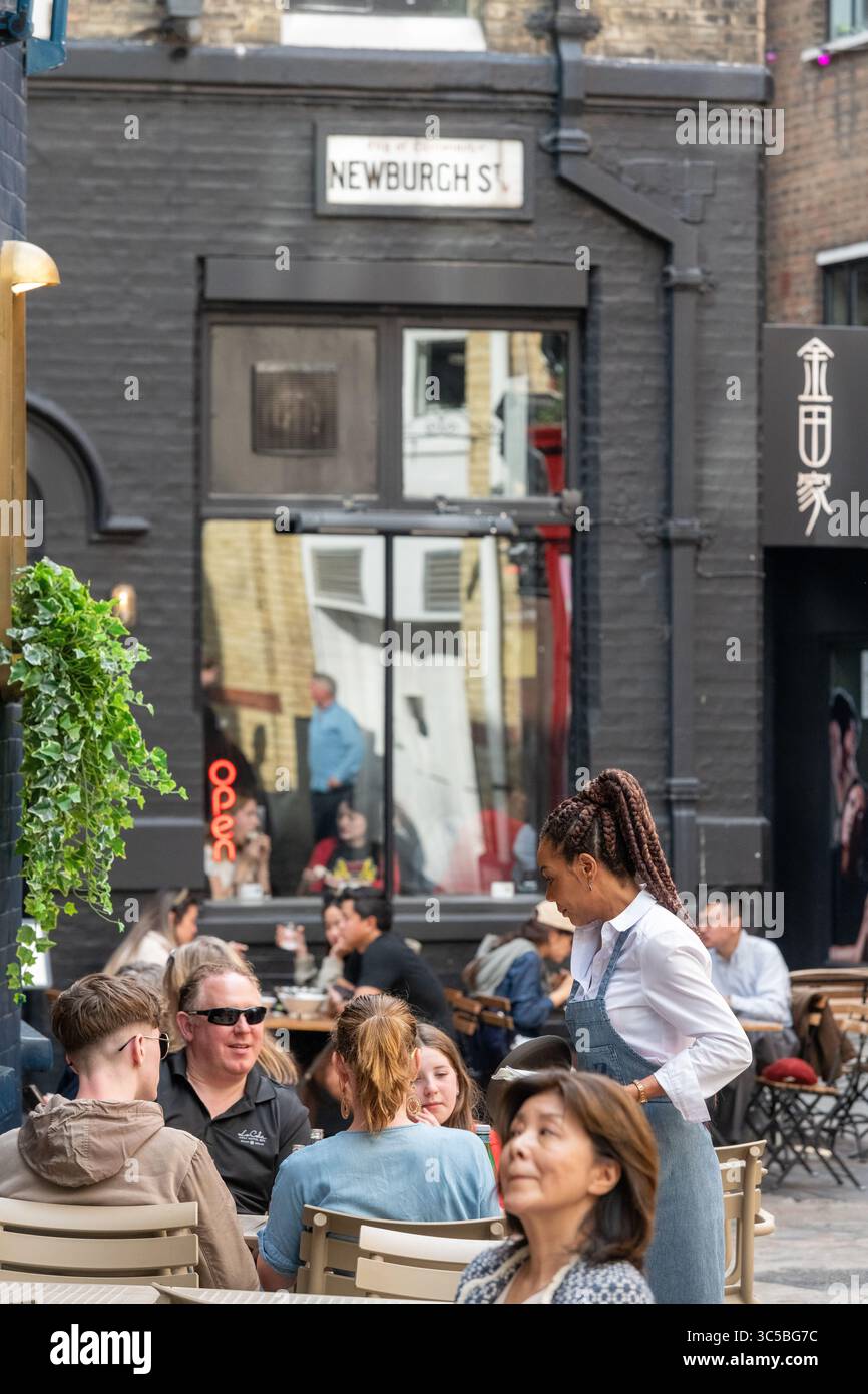 Busy outdoor dining scene on Newburgh Street in Soho, London, with diverse people enjoying al fresco food and drinks on a warm summer day. Stock Photo