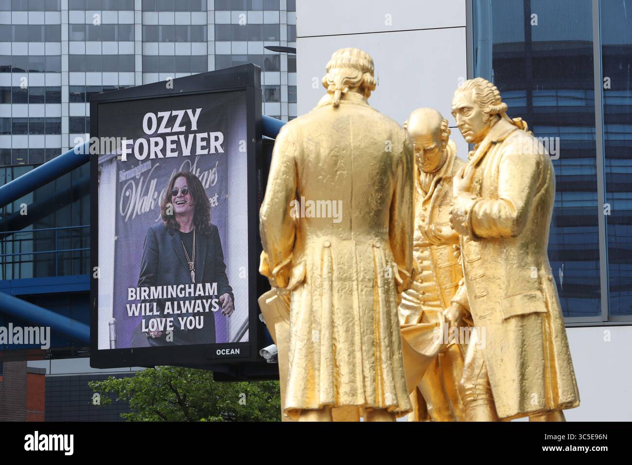 Birmingham, UK. 30th July, 2025. Thousands of rock fans have arrived in Broad Street, Birmingham, to see the cortege of the late rock star Ozzy Osbourne as it makes its way to Black Sabbath Bridge. The digital screen is seen next to a statue of past Birmingham heroes Boulton, Watt and Murdoch, nicknamed 'The Golden Boys'. Credit: Alamy Live News Stock Photo