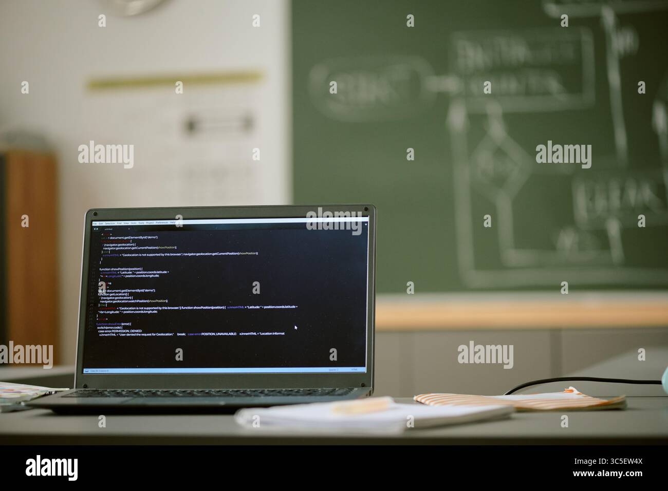 Laptop Displaying Programming Code in Classroom with Chalkboard in Background Stock Photo