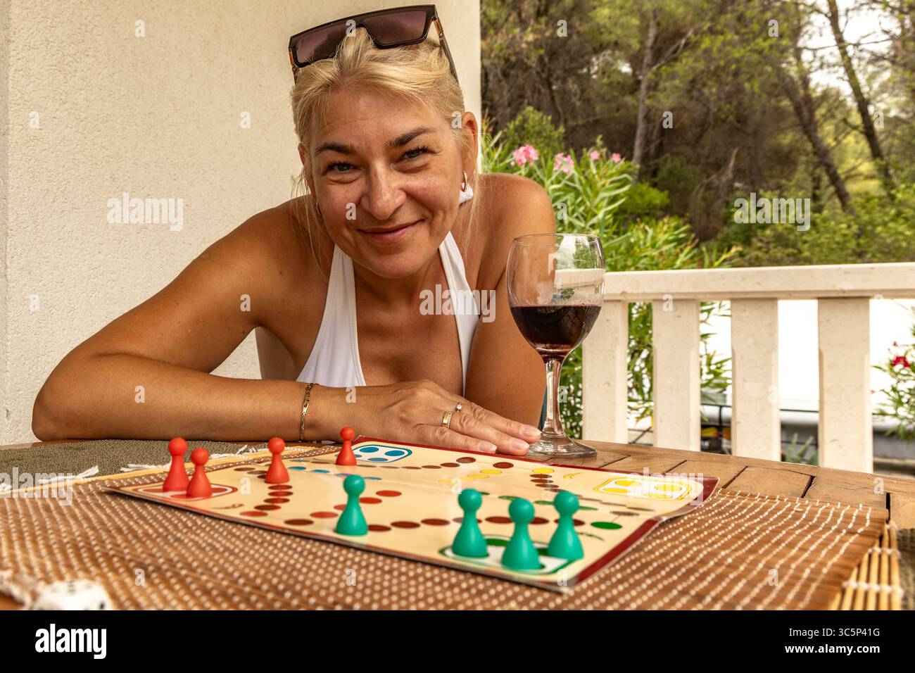 A woman with a glass of red wine sits at a table and plays a board game, playing Chinese checkers, a way to spend free time on vacation Stock Photo
