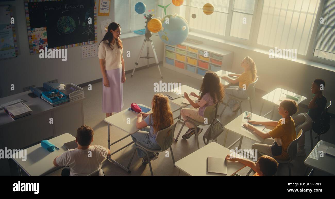Female Teacher Finishing Lesson, Announcing the End of School Day to Diverse Kids. Elementary School Students Standing Up, Packing Their Backpacks and Leaving Modern Classroom. Slow Motion. High Angle Stock Photo