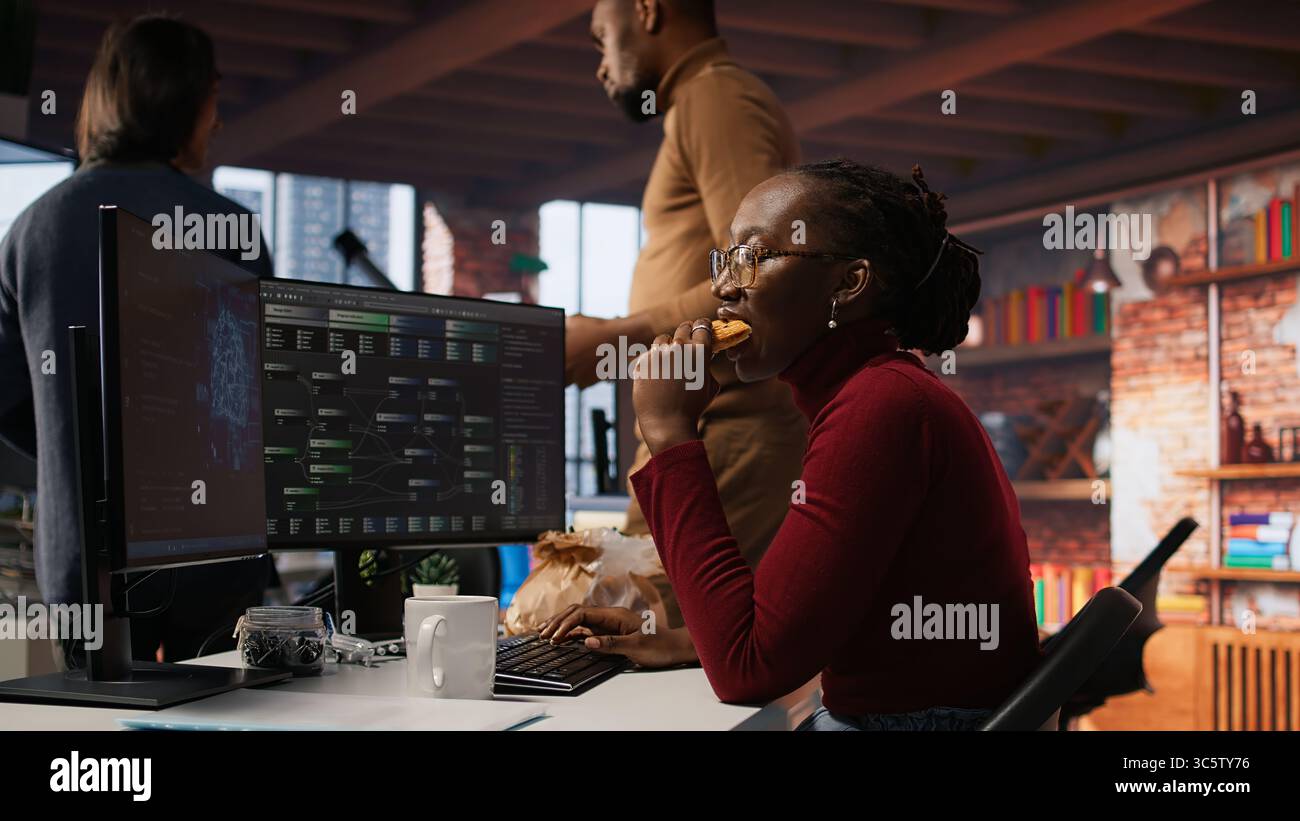 Programmer coding AI algorithm in startup, eating food at desk while refining machine learning models. Stressed woman using coding platform in Silicon Valley office and enjoying snack, camera A Stock Photo