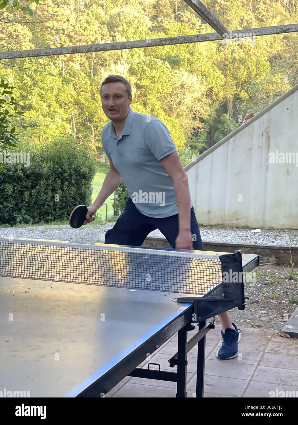 Table tennis enthusiast engaged in a lively backyard game. The player, focused and energized, strikes the ball with determination, surrounded by a lus Stock Photo