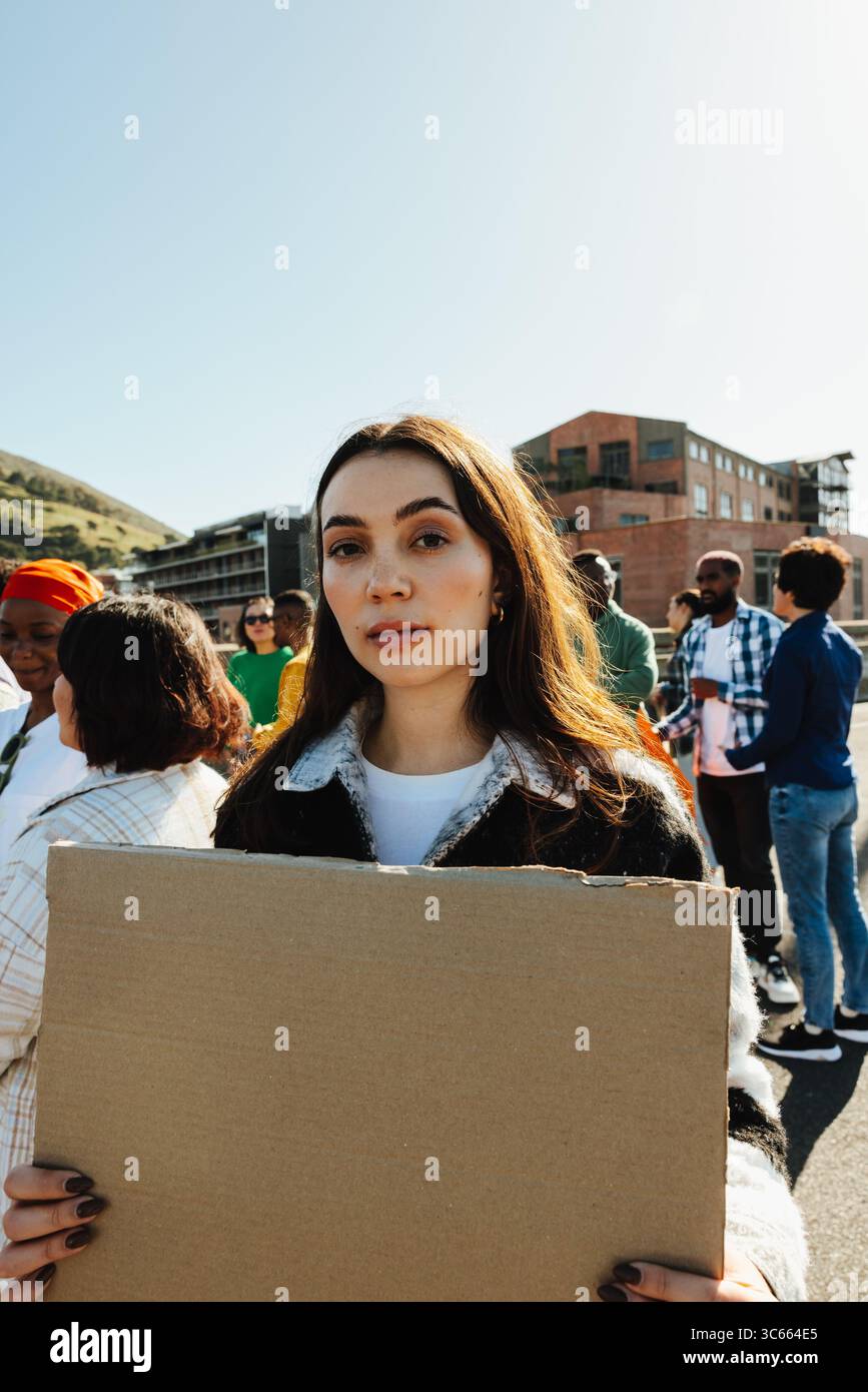 A young woman stands outdoors holding a blank sign, surrounded by a diverse group of people demonstratively gathered on a sunny day, symbolizing activ Stock Photo