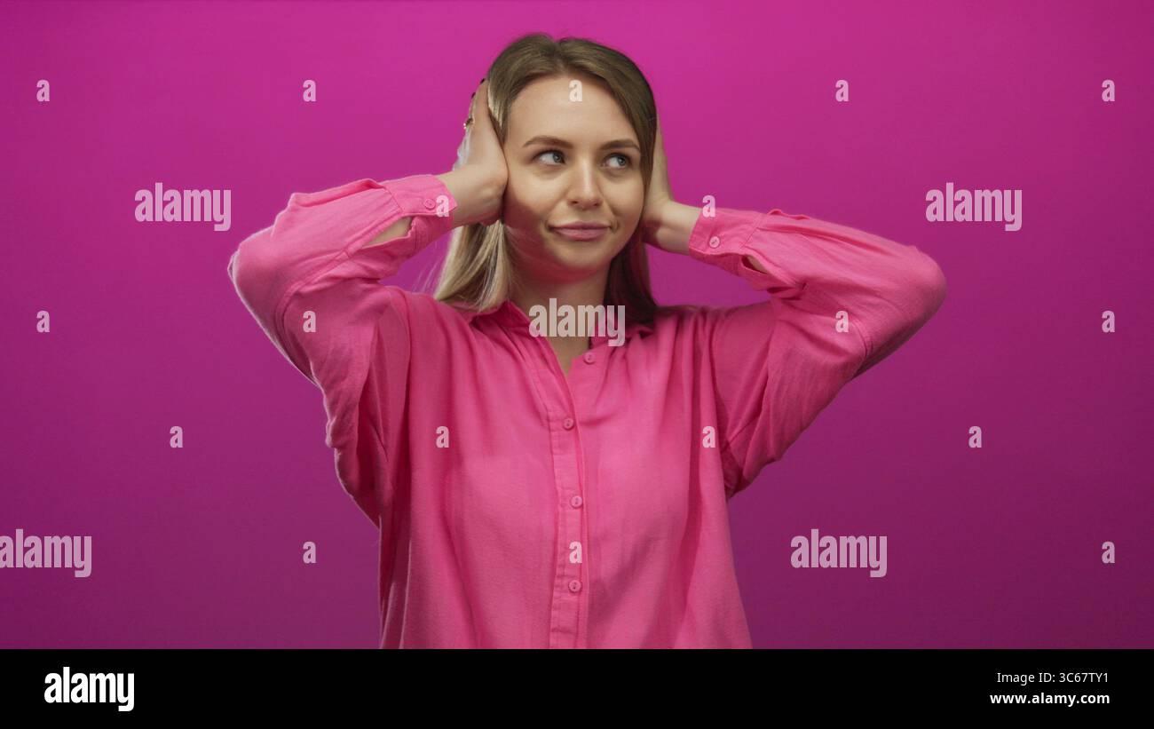 Young woman in pink shirt covers ears with hands, expressing frustration against a vivid pink background, conveying a sense of isolation and emotional Stock Photo